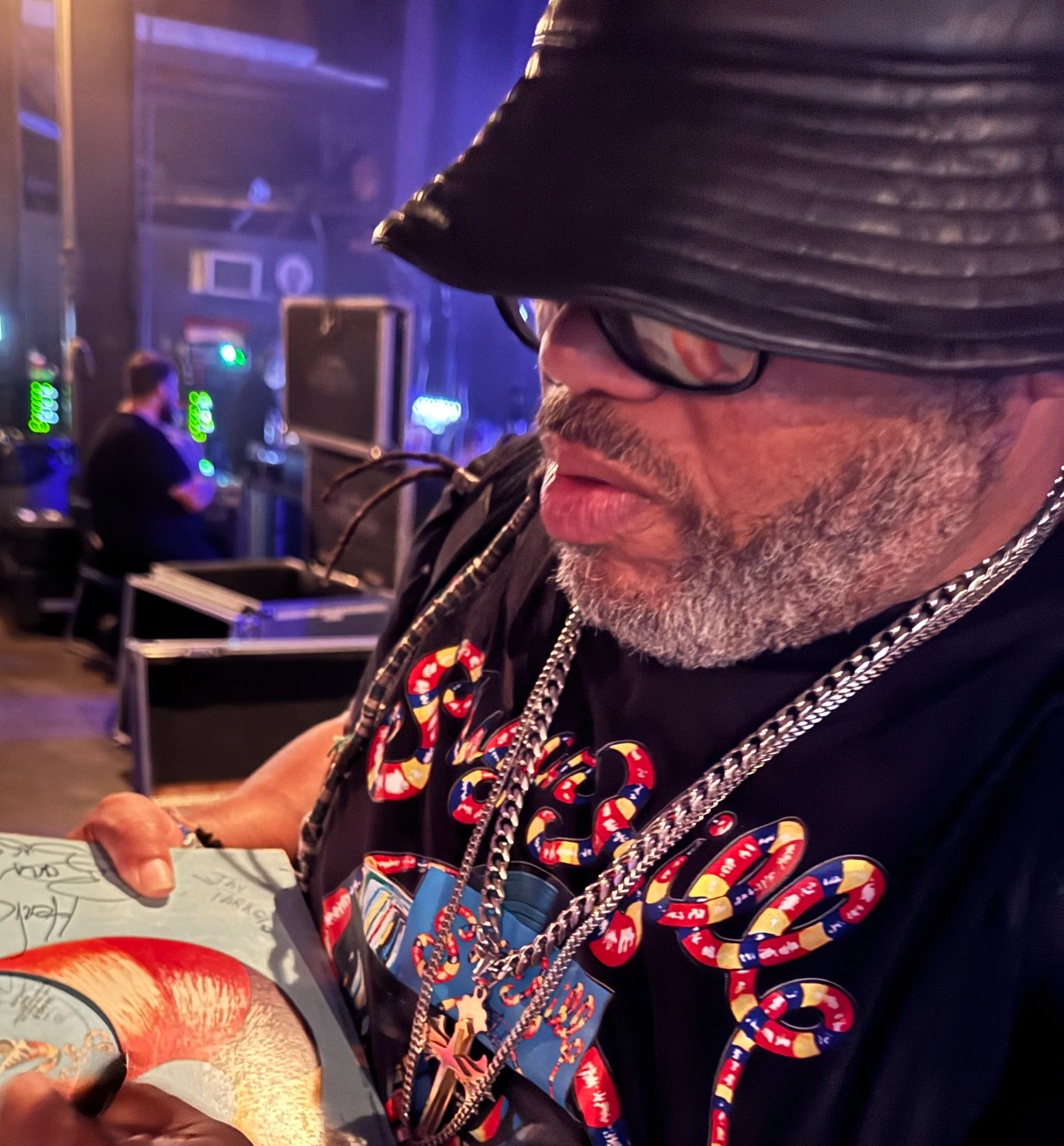 A man with gray facial hair, wearing glasses, a black leather hat, and a black t-shirt with colorful design, is signing for a fan in a dimly lit room with blue and purple lighting, and in the background, a person is sitting at a table.