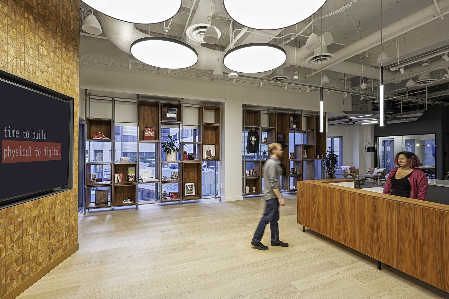 Modern office lobby with a wooden reception desk, shelving with books and decorations, large circular ceiling lights, and a seating area with sofas and chairs. Two women are present, one at the reception and the other walking past.