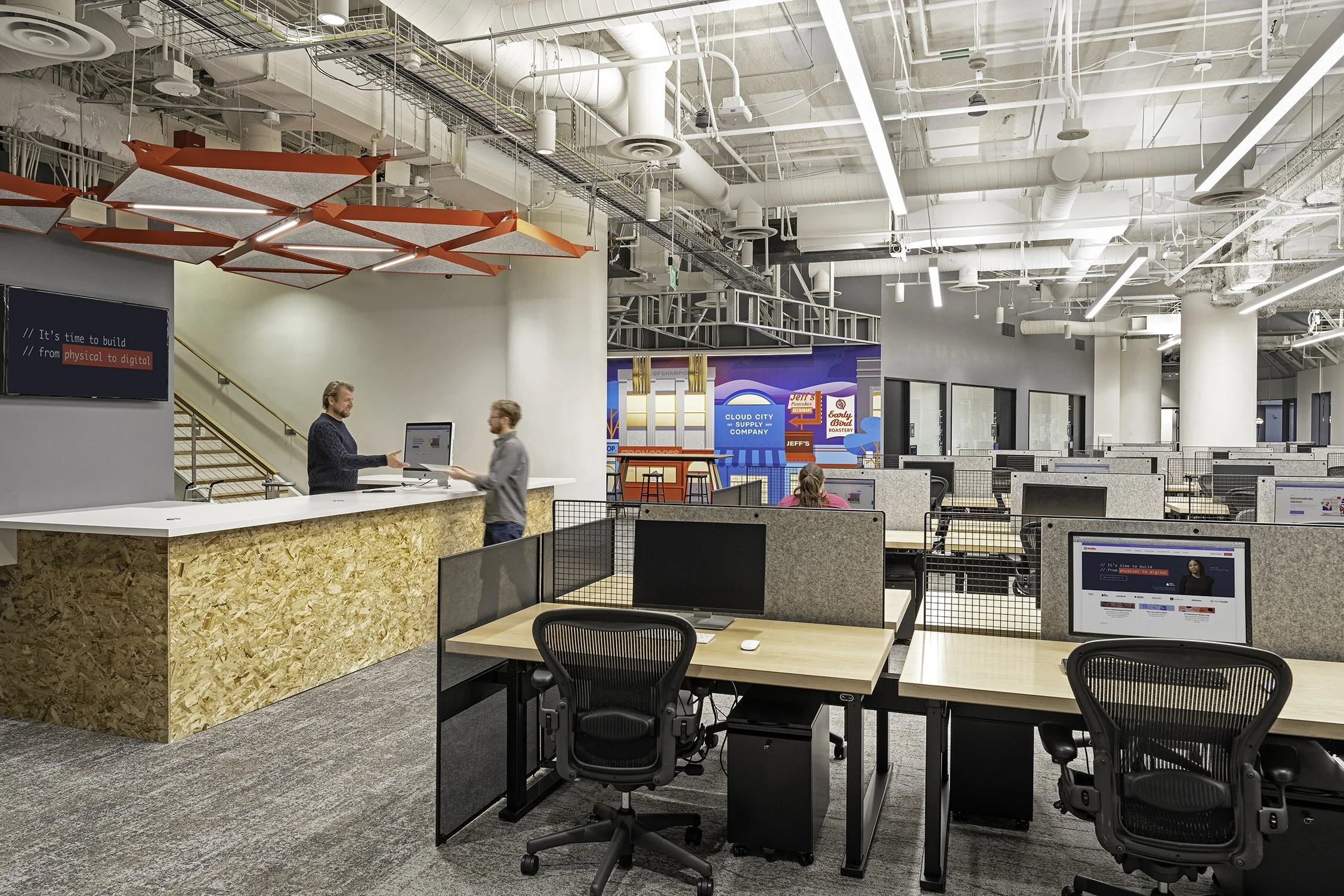 Open coworking space with rows of desks and computers, a reception counter with two people, modern lighting, large digital screens, and a vibrant mural.