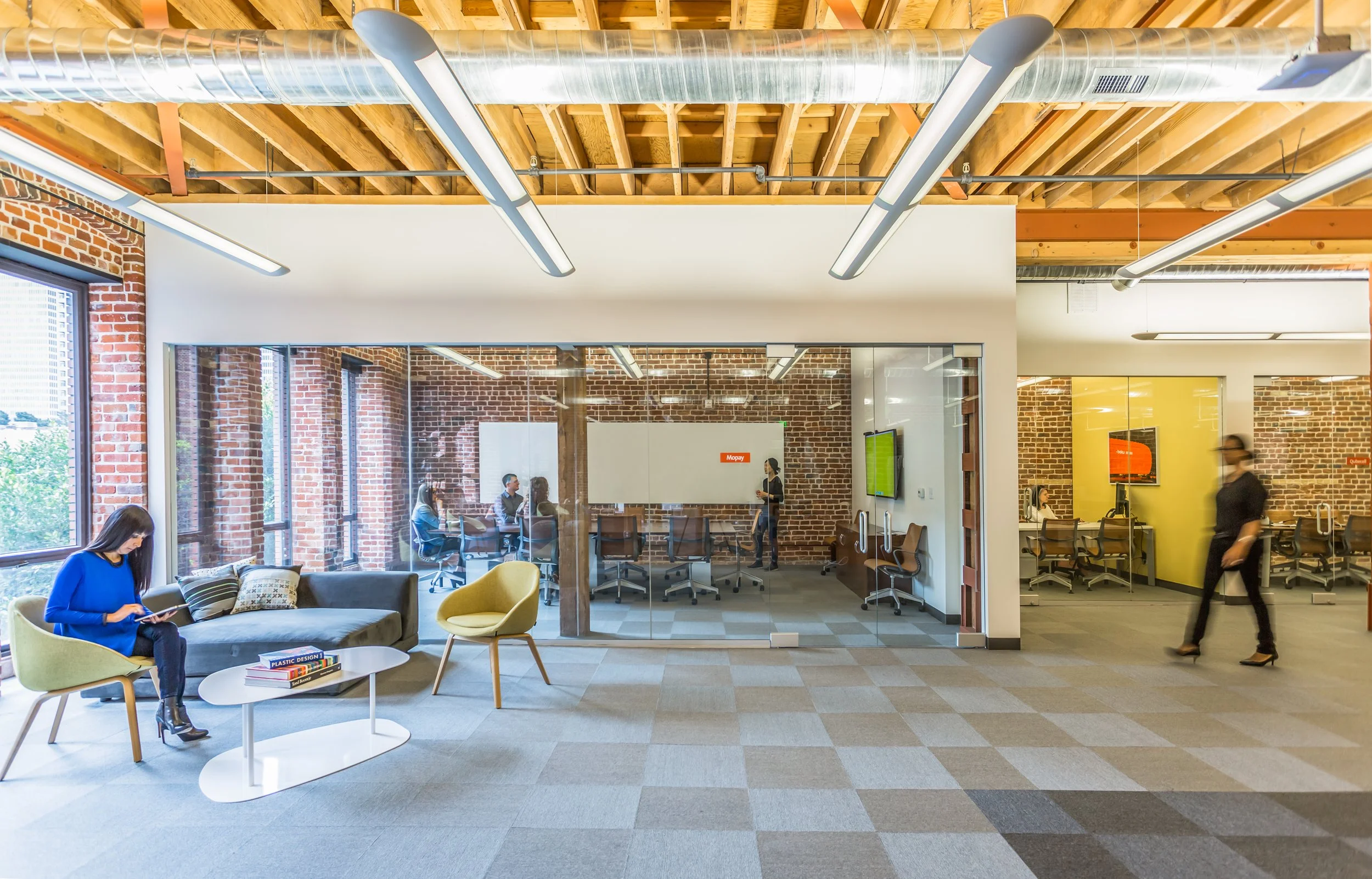 Modern open office space with exposed brick walls, large windows, and wood ceiling beams. There are people working and walking, some sitting at conference tables and others walking through the area.