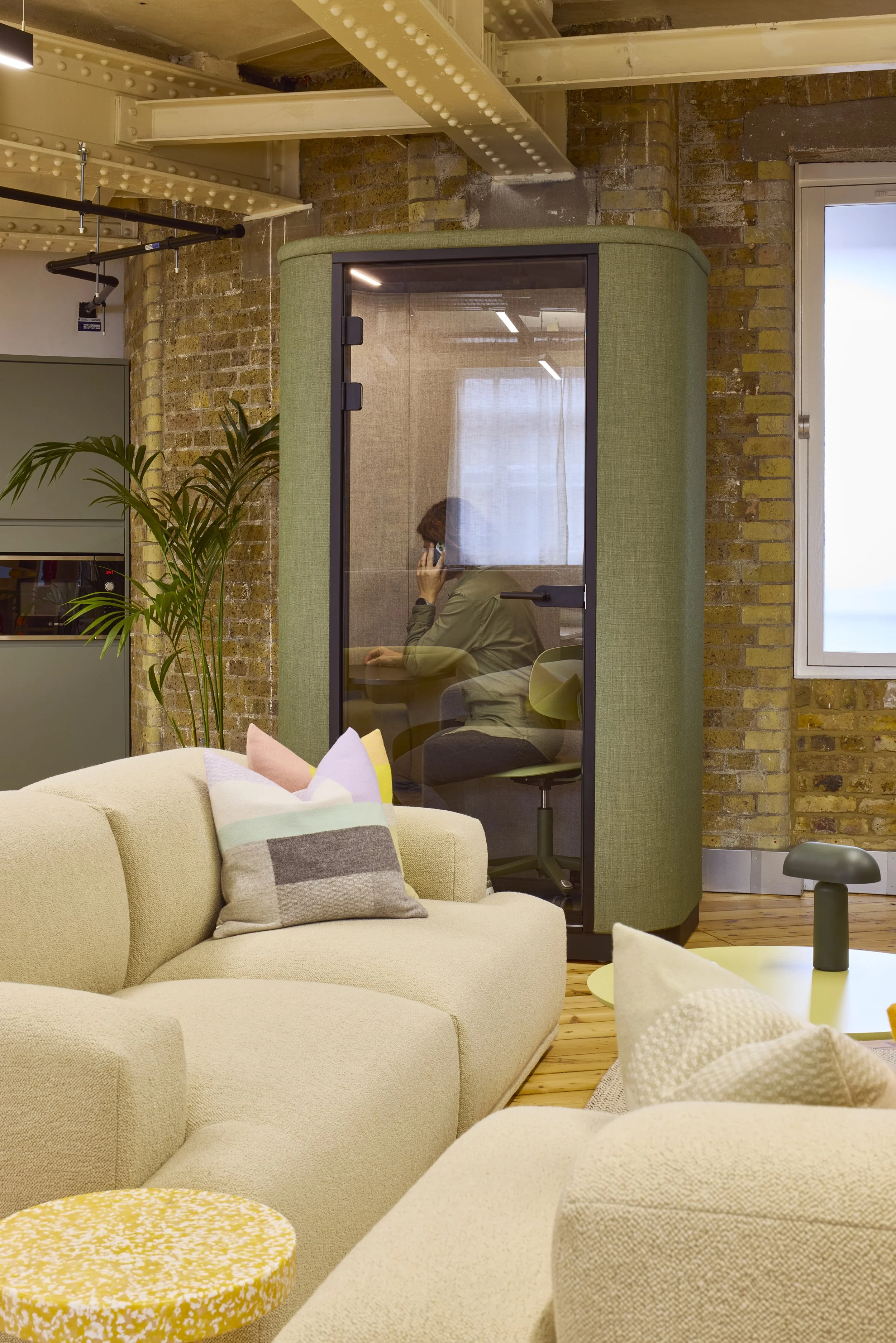 A woman seated inside a soundproof booth talking on a phone in an office space with brick walls, a window, and modern furniture.