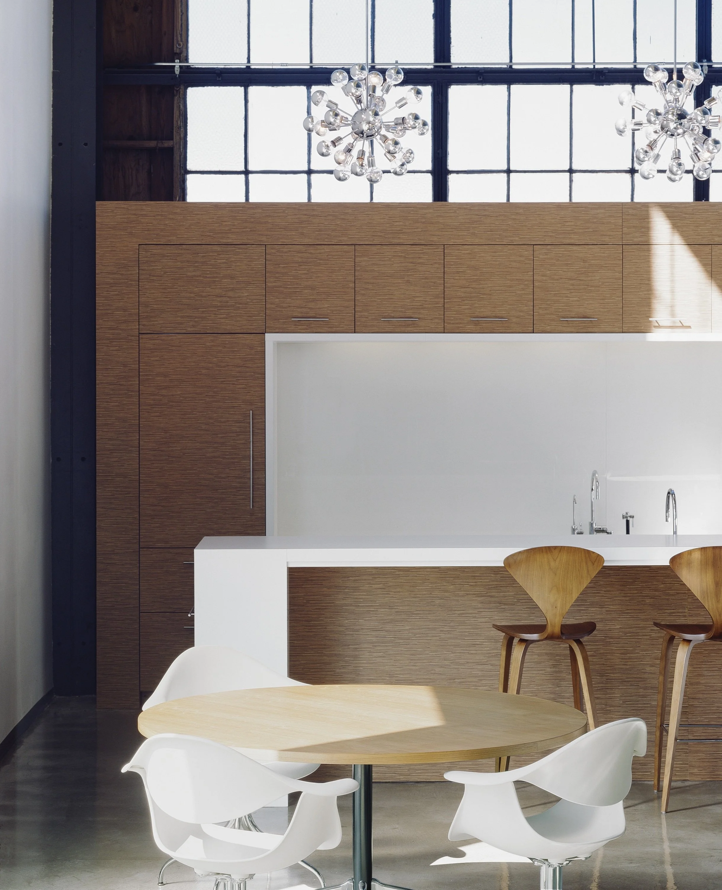 Modern kitchen area with wooden cabinetry, a white countertop, and wooden barstools, featuring large industrial-style windows and contemporary chandeliers hanging from the ceiling.