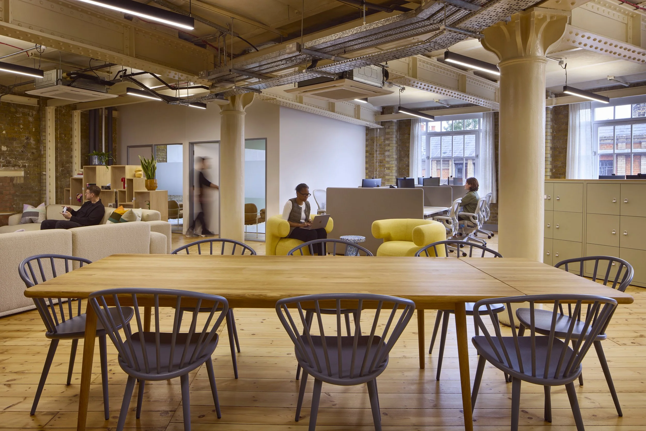 Open-plan coworking space with a large wooden table surrounded by black chairs in the foreground, and seating areas with beige sofas and yellow armchairs in the background. There are people working on laptops and using their phones, with natural light coming through large windows.