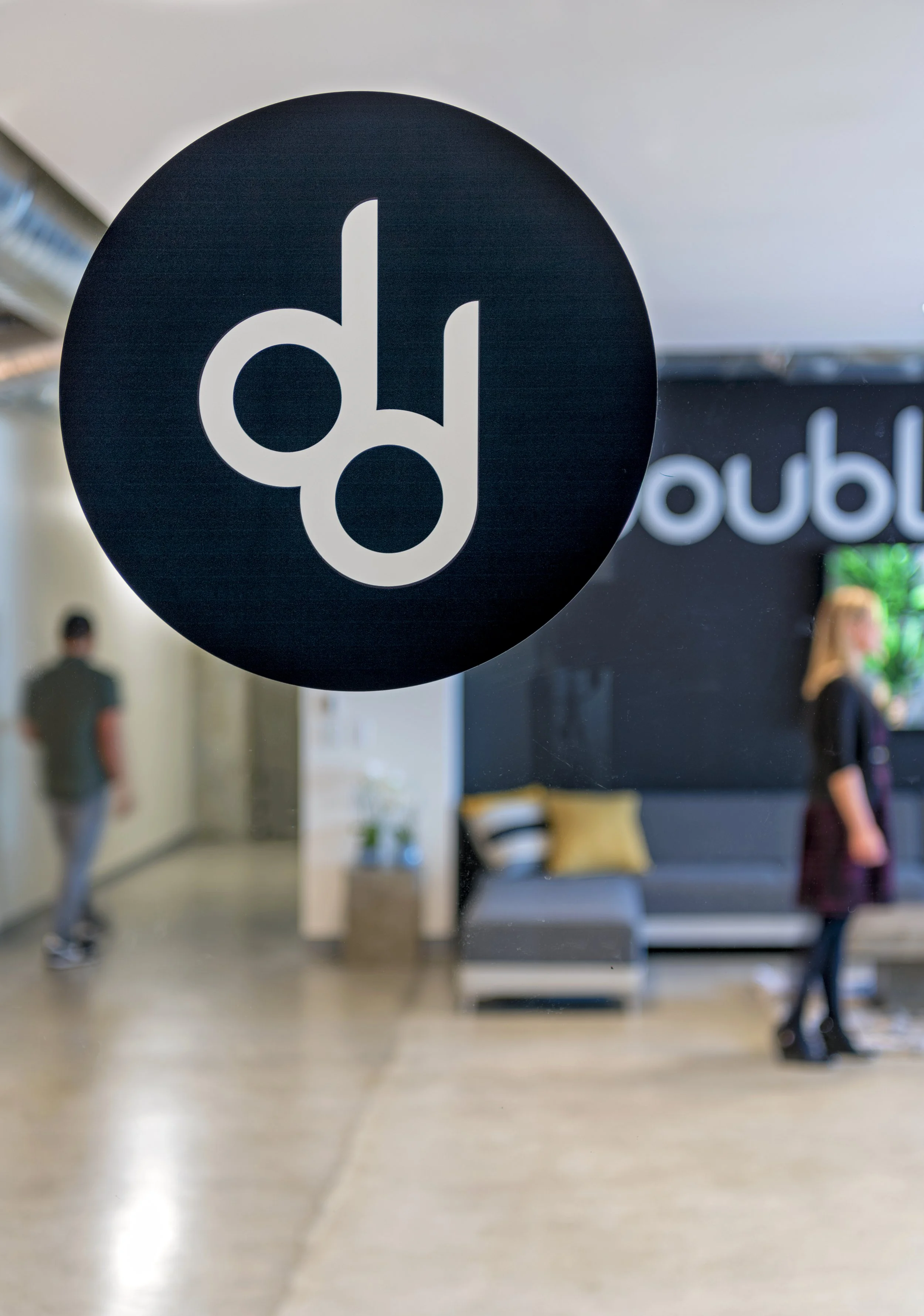 Close-up of a round black sign with white lowercase 'd' and 'd' letters, hanging in an indoor space with blurred background of people and a reception area.