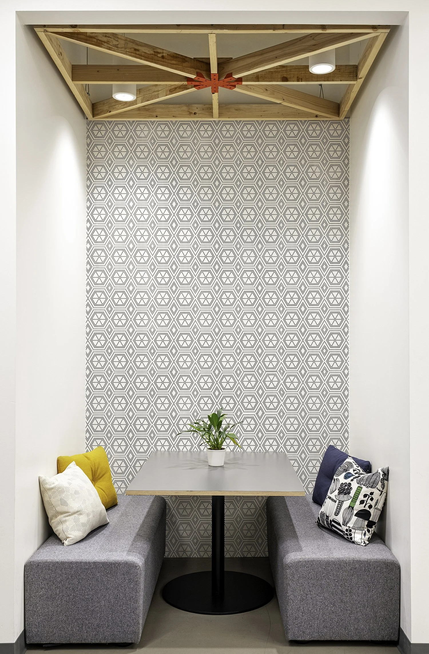 Small cozy dining nook with a table, built-in benches with colorful pillows, a geometric patterned accent wall, a potted plant at the center of the table, and a decorative wooden ceiling structure with two ceiling lights.