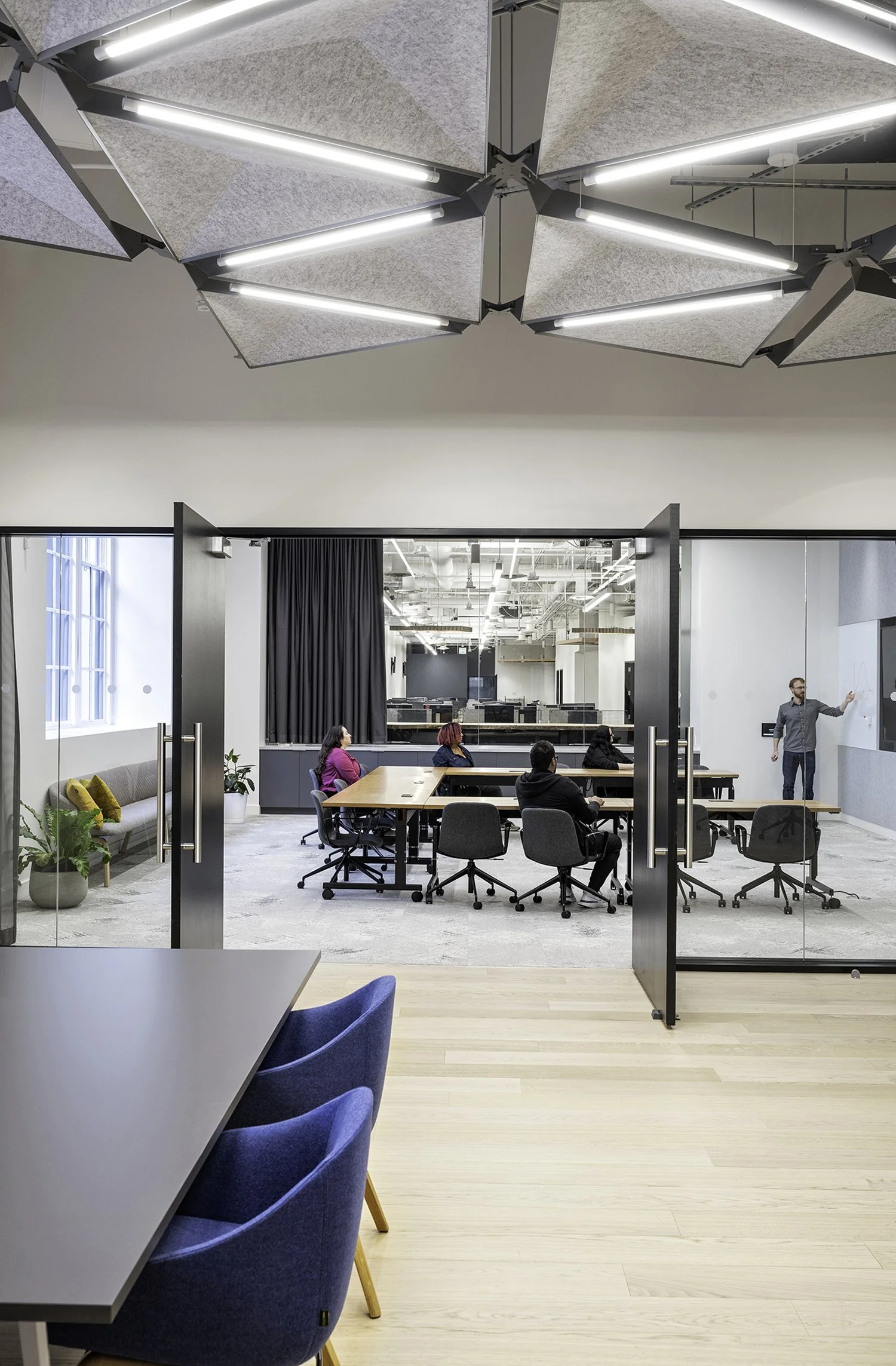 A modern conference room with four people seated at a rectangular table, listening to a presenter standing at a whiteboard, seen through glass doors from an adjacent room with a blue chair and a table.