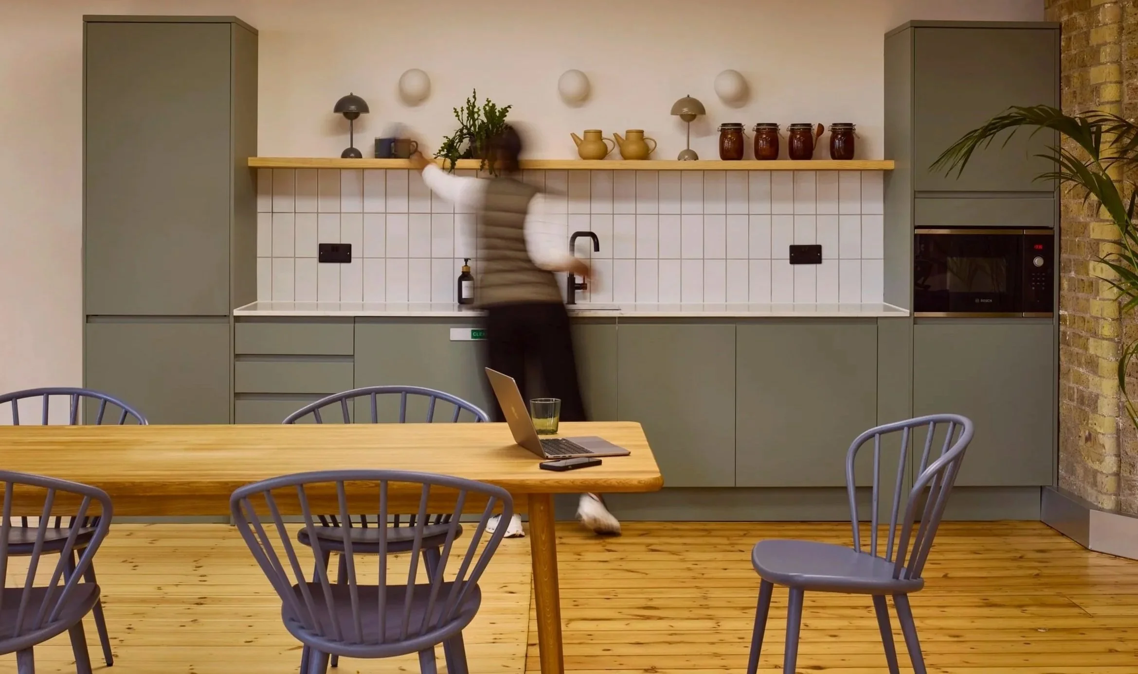 Modern kitchen with green cabinets, white tiled backsplash, and wooden shelves. A person is reaching for items on the shelf, with a laptop and a glass of water on the wooden dining table in the foreground.