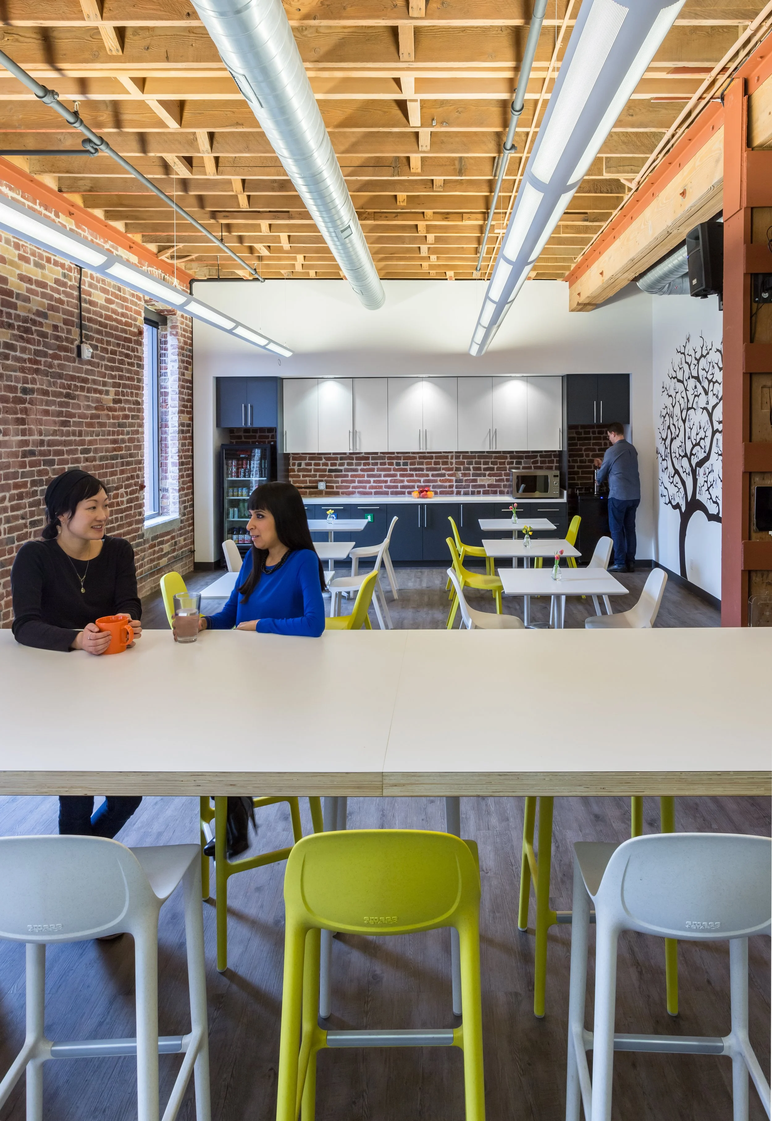Two women seated at a white counter drinking coffee and chatting in a modern, industrial-style cafe with brick walls, exposed wooden ceiling beams, and a kitchen area with white cabinets and a brick backsplash in the background.