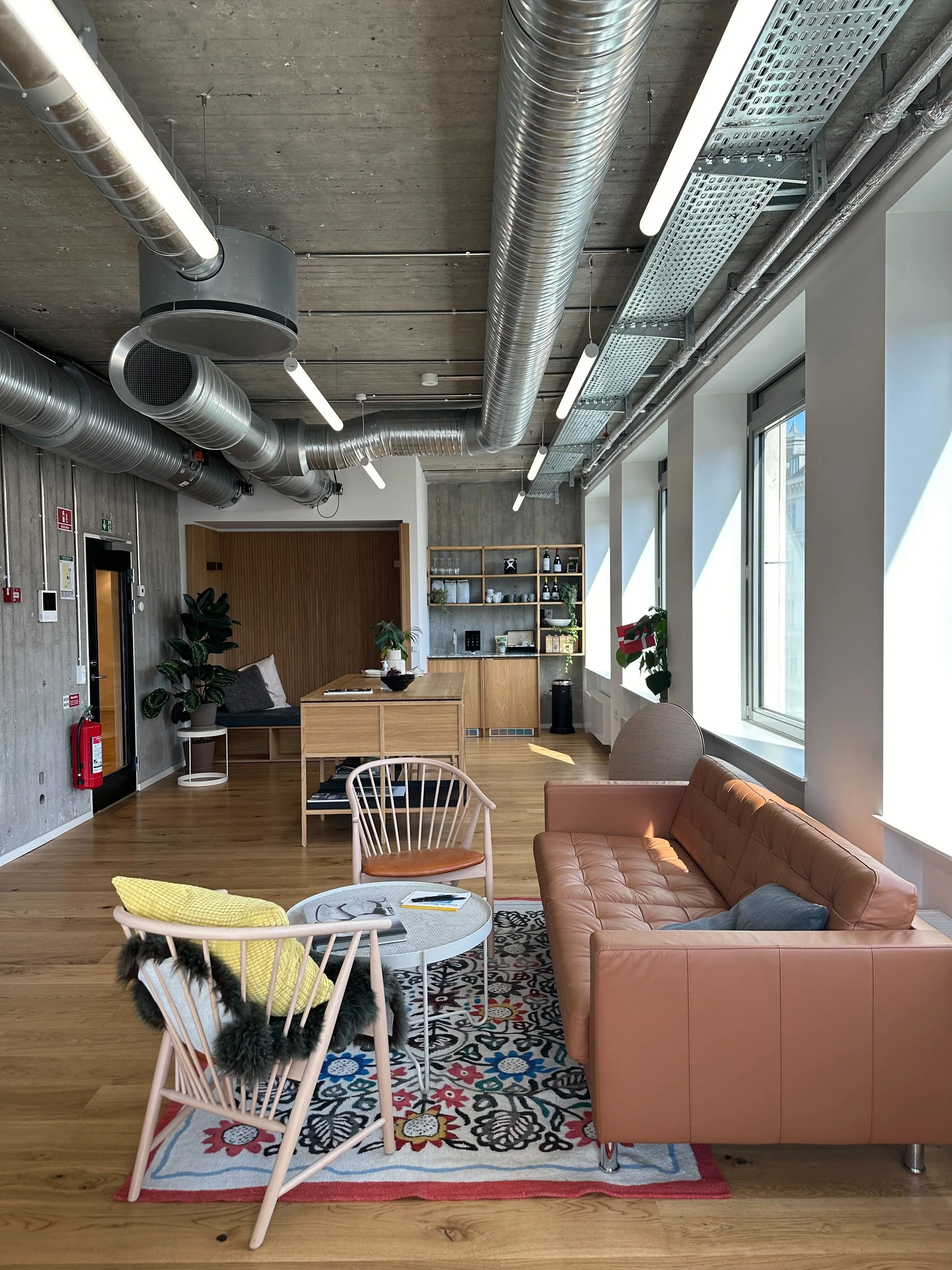 Modern office lounge with wooden floors, a peach leather sofa, two light pink chairs, a small round table with a yellow pillow and fluffy black blanket, and large windows letting in natural light. There are plants, a bookshelf, and exposed ductwork on the ceiling.