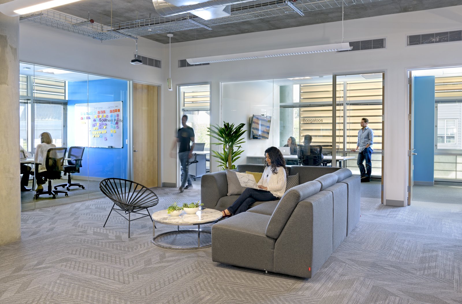 Modern office lounge with a woman sitting on a gray sofa working on a laptop, a round coffee table with plants, glass-walled meeting rooms in background with people working, and a large plant in the center.