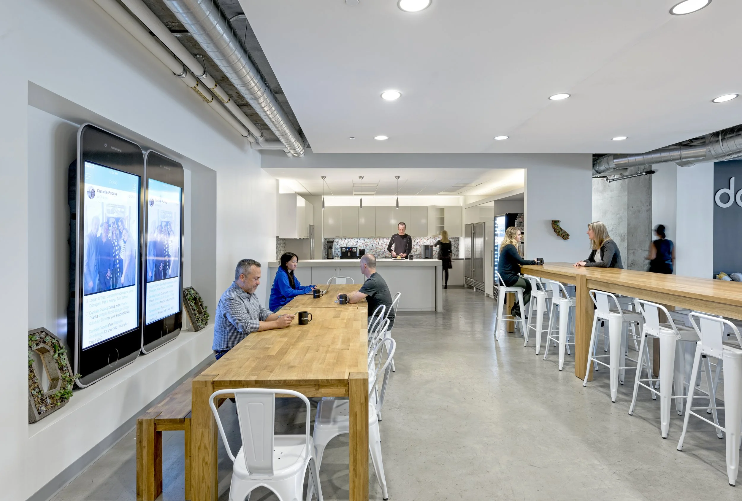 Modern cafe or coworking space with high tables, bar stools, and a kitchen in the background. People are sitting at tables, some drinking coffee and working on laptops. Large digital screens on the left wall display social media posts.