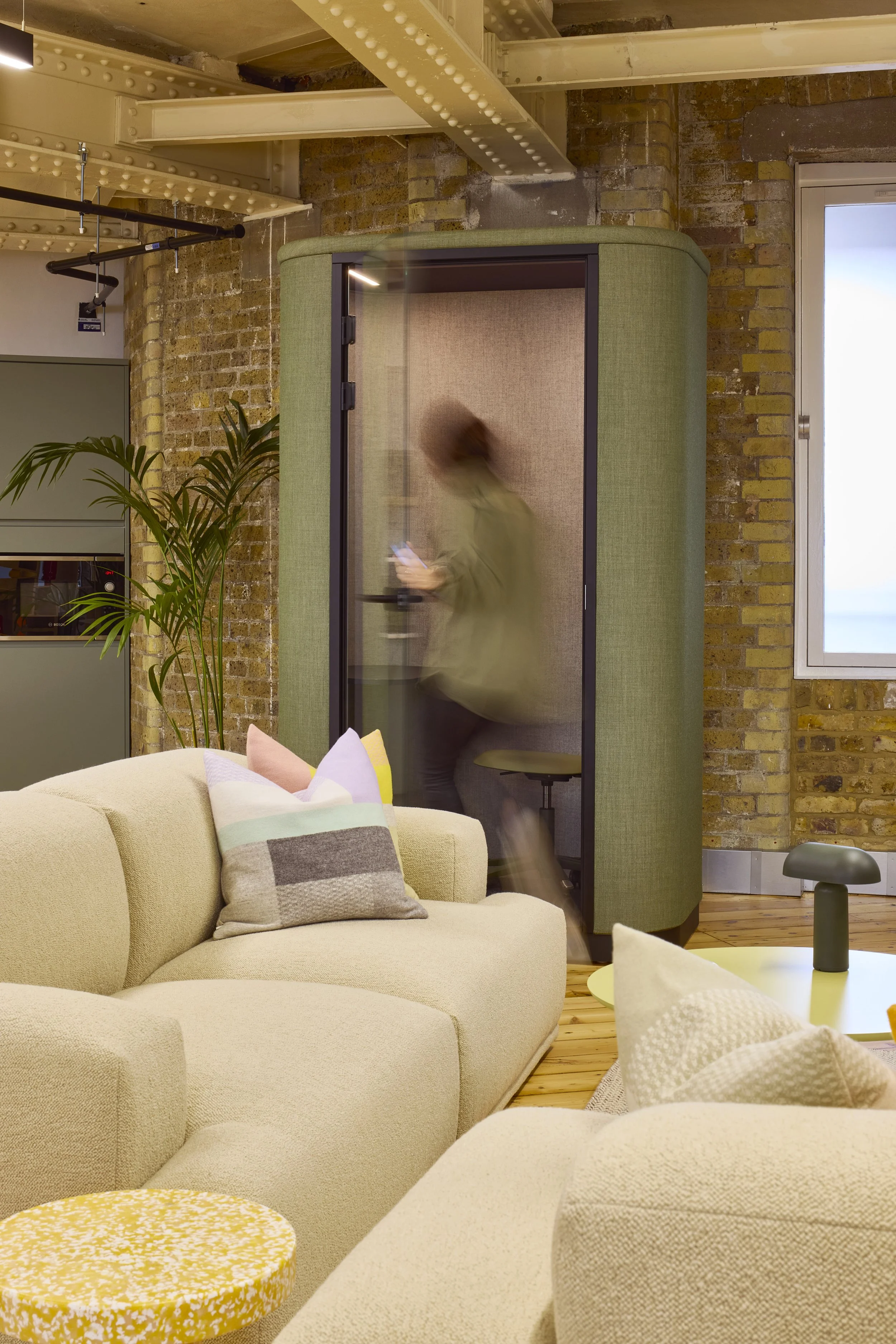 A woman walking through a modern office space, blurred as she looks at her phone, with beige sofas and colorful pillows in the foreground, a green plant, brick wall, and window in the background.