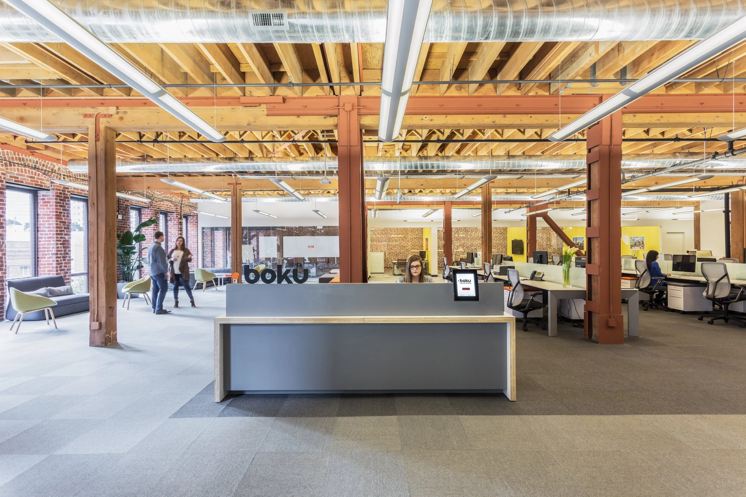 Office reception area with a gray front desk, a woman working on a computer, an open workspace with multiple desks and chairs, a few people walking and talking, and large windows letting in natural light, with exposed brick walls and wooden beams on the ceiling.