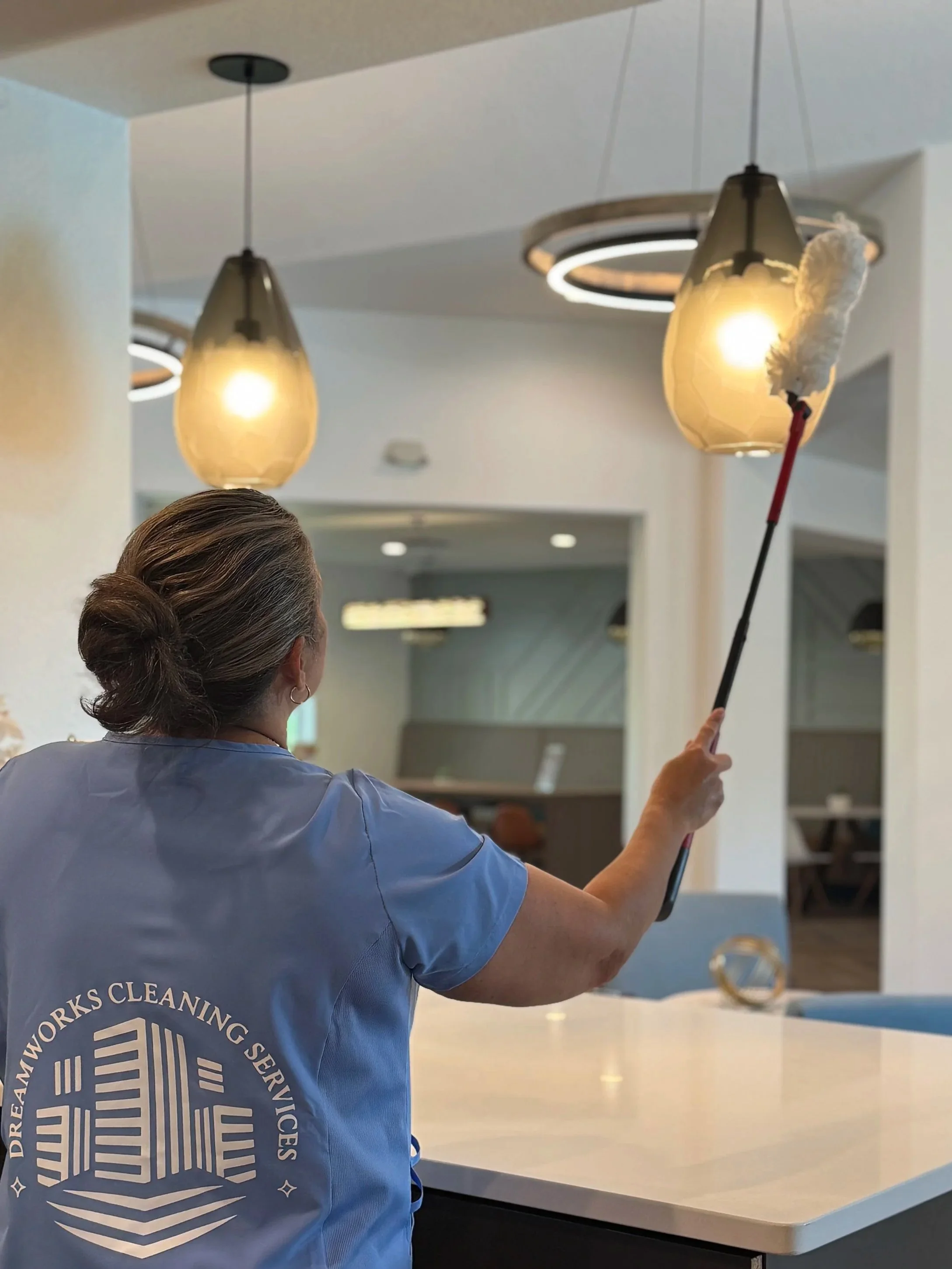 A cleaning staff member wearing a blue uniform with DreamWorks Cleaning Services logo is dusting a ceiling light fixture with a long duster in a modern, well-lit interior space.