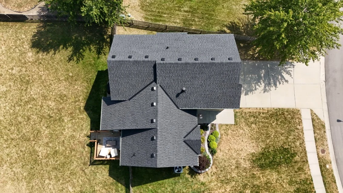 Top-down aerial view of a house with gray roof shingles, surrounded by a lawn, trees, and a sidewalk, with a driveway on the right side.