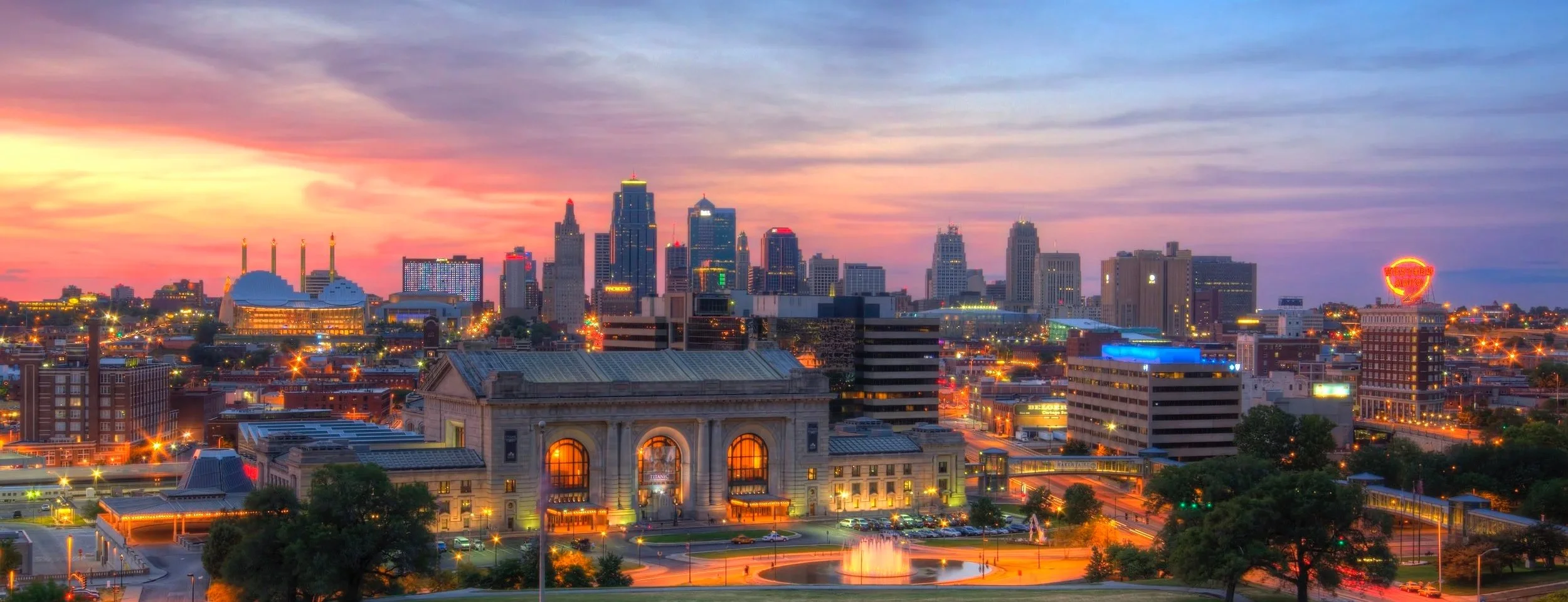 Sunset over a city skyline with tall buildings, a historic train station, and vibrant street lights.
