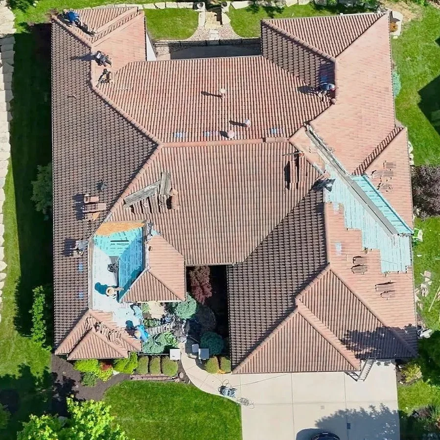 An aerial view of a house with a complex roof structure covered in reddish-brown tiles. The roof has multiple sections, some under construction or repair, with workers visible. The house is surrounded by a green lawn, a driveway, and shrubs.