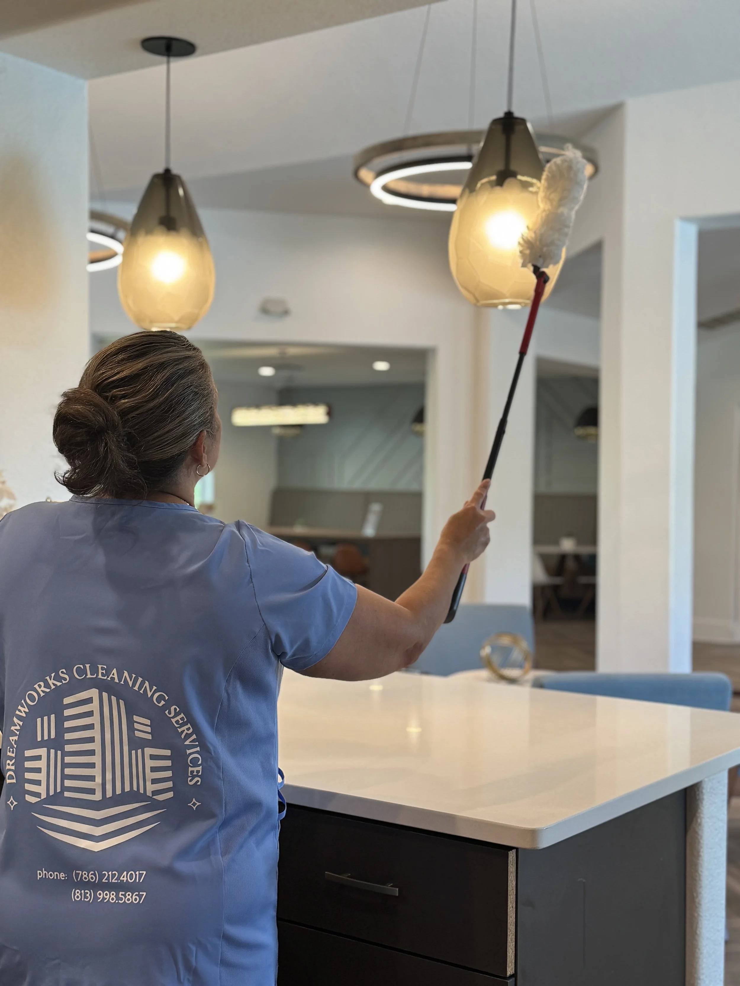 A housekeeping staff in a blue uniform cleaning a white counter with a duster on a long-handled tool in a modern interior space.
