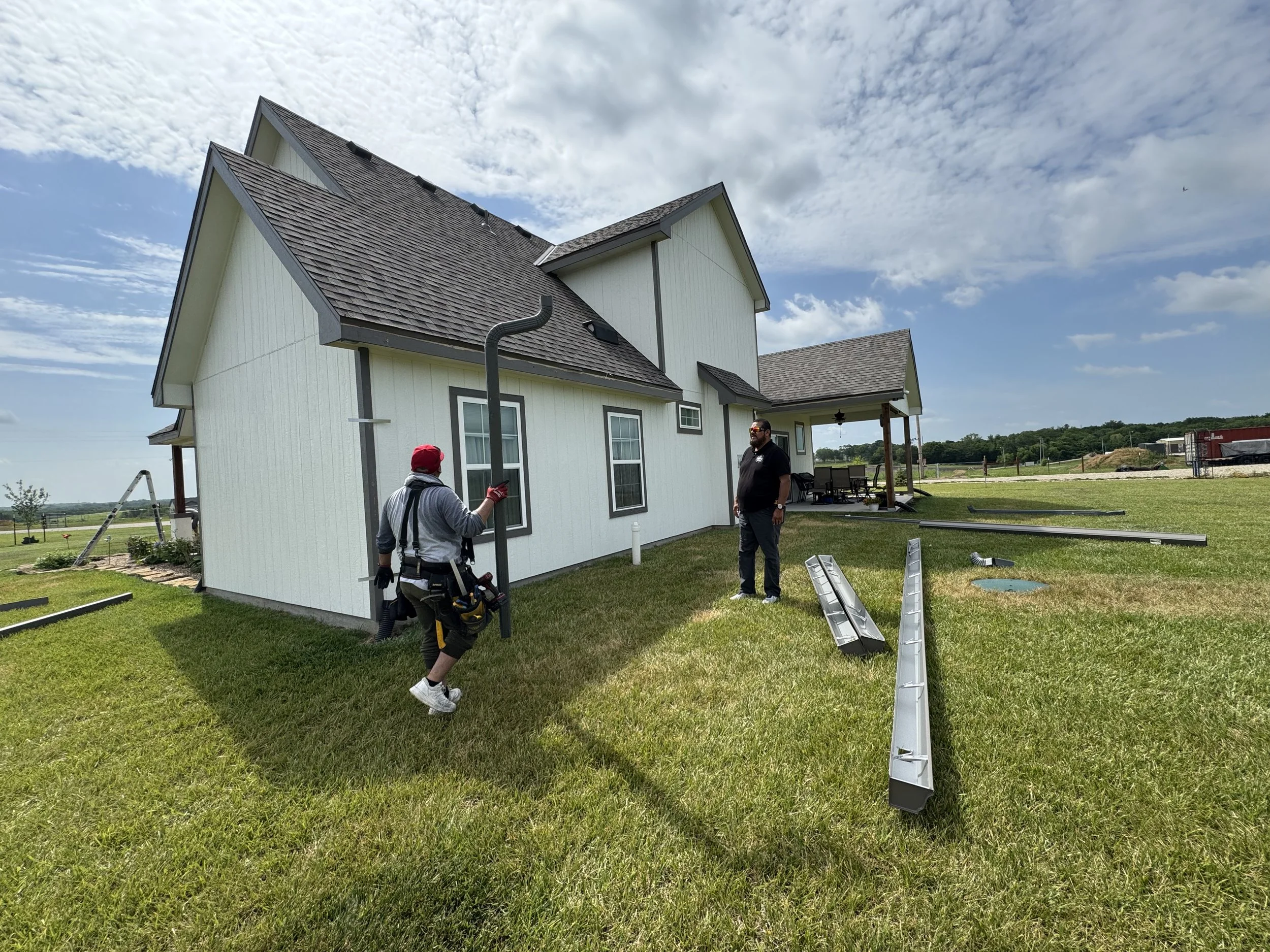 Construction workers installing a gutter on a white two-story house with black trim, on a sunny day with partly cloudy sky.