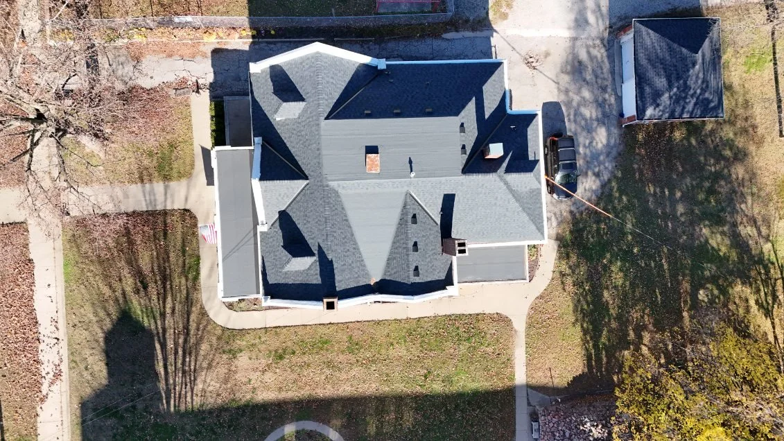 Aerial view of a large house with grey roof and surrounding yard, driveway, and trees.