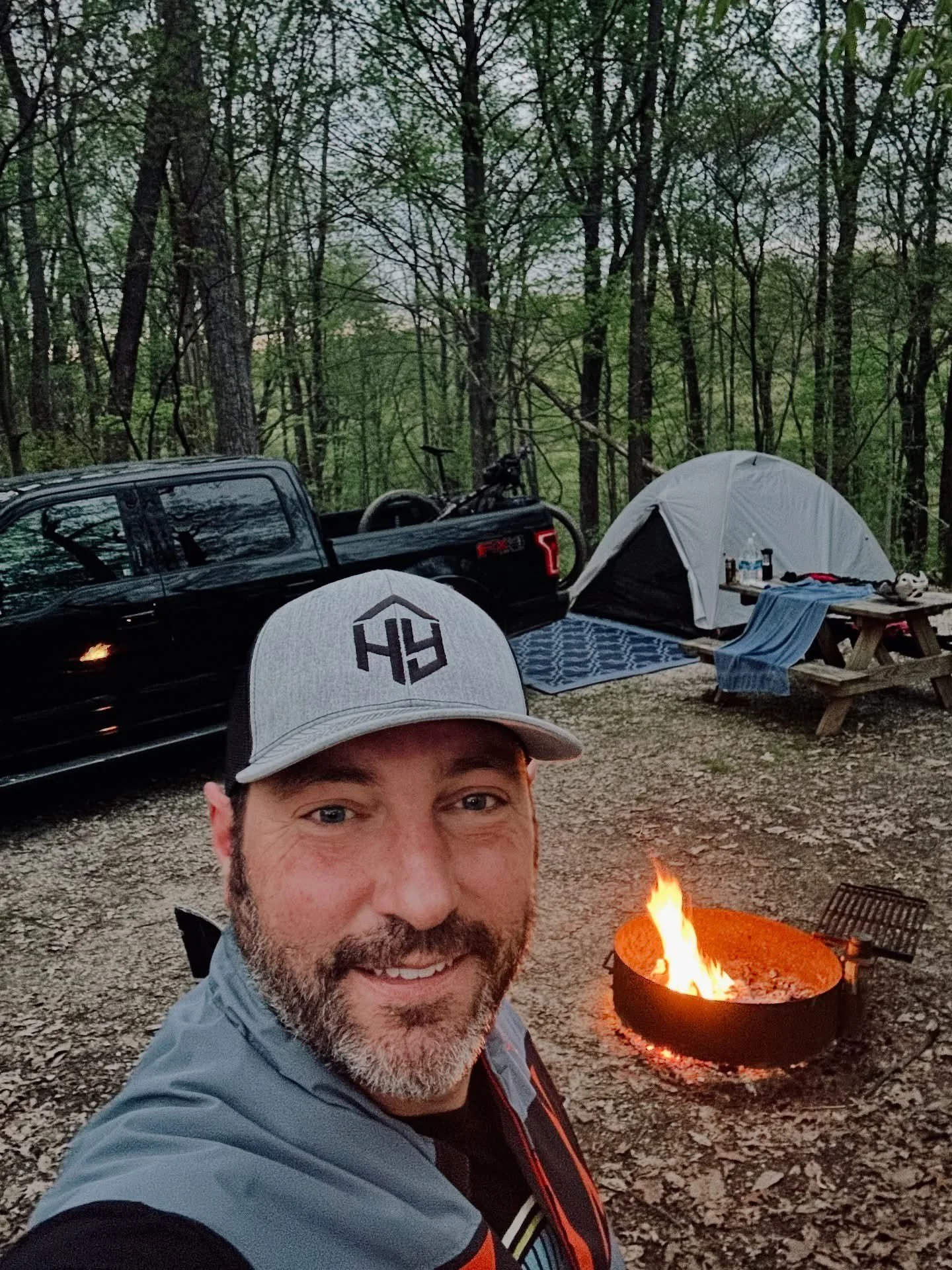 A man smiling with a camping site in the background, including a tent, a truck, a picnic table with items on it, and a campfire in a wooded area.