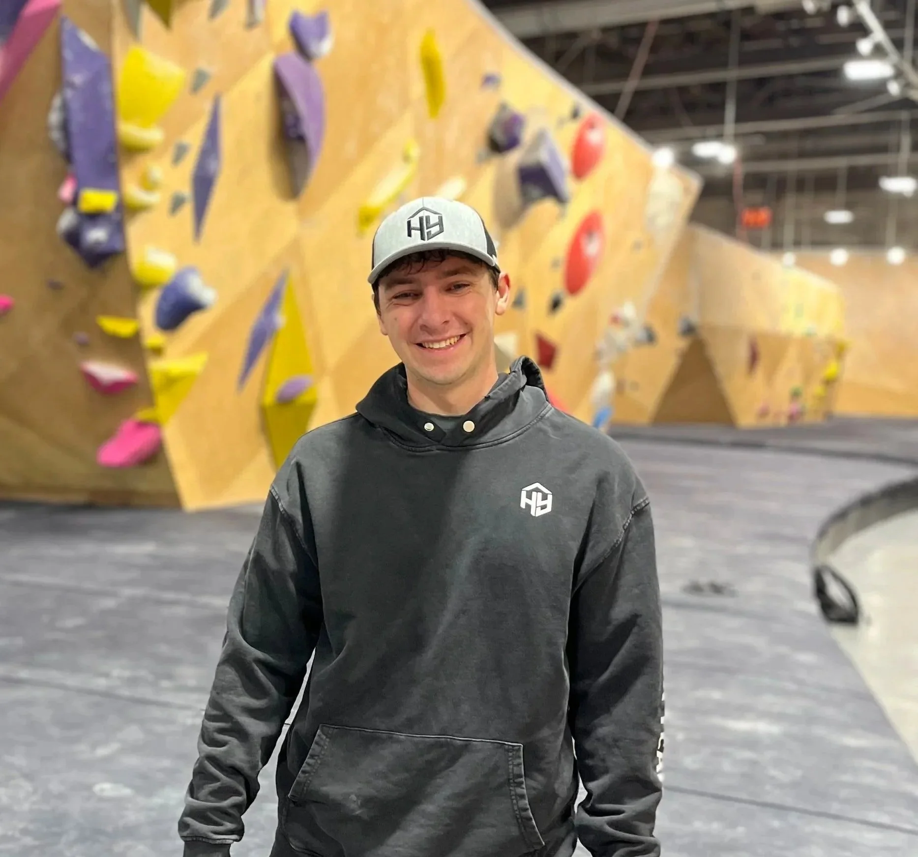 Young man smiling at an indoor climbing gym, wearing a gray baseball cap and a dark gray hoodie, with colorful climbing holds in the background.