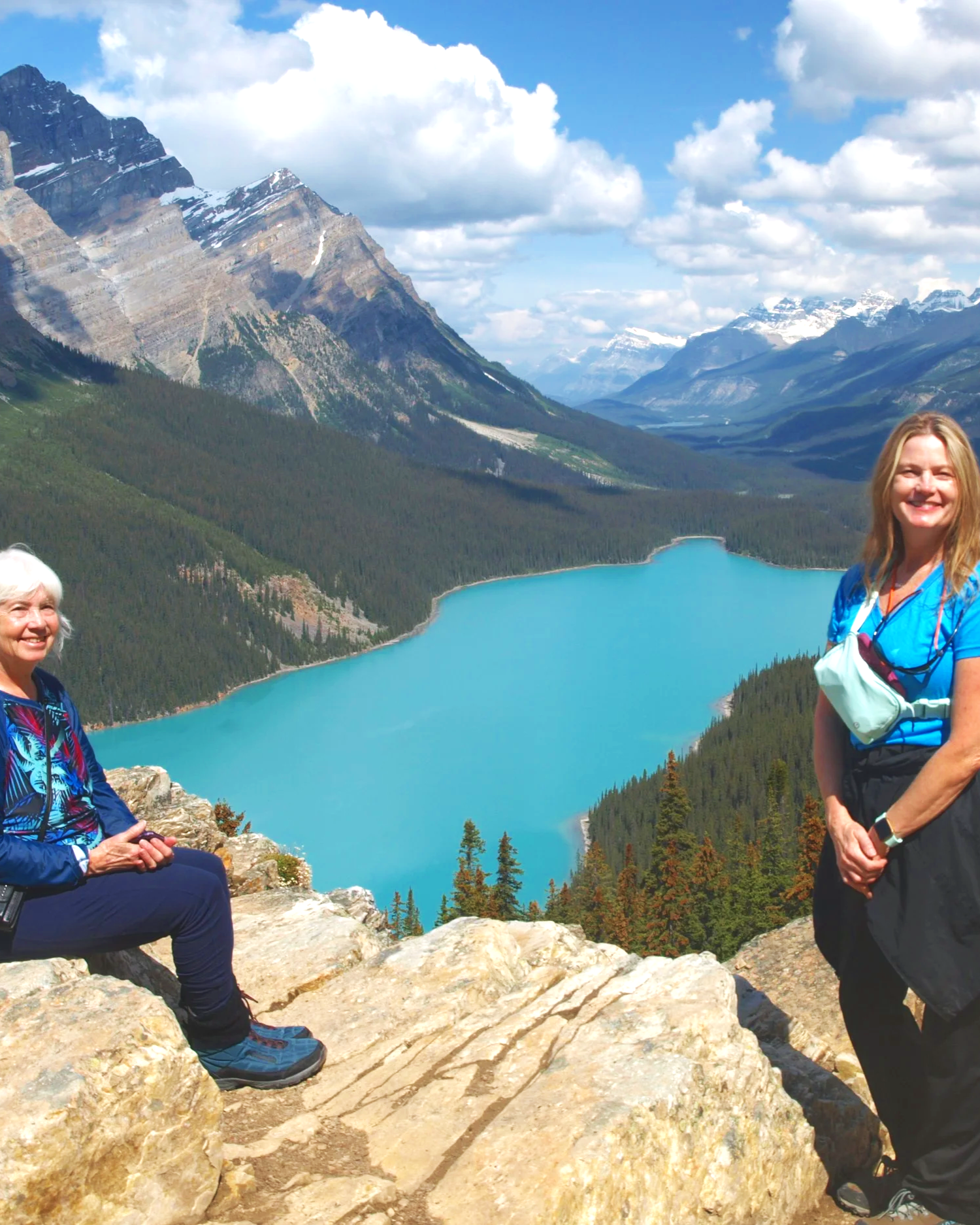 Two women on a rocky ledge overlooking a turquoise lake surrounded by forest and mountains under a partly cloudy sky.