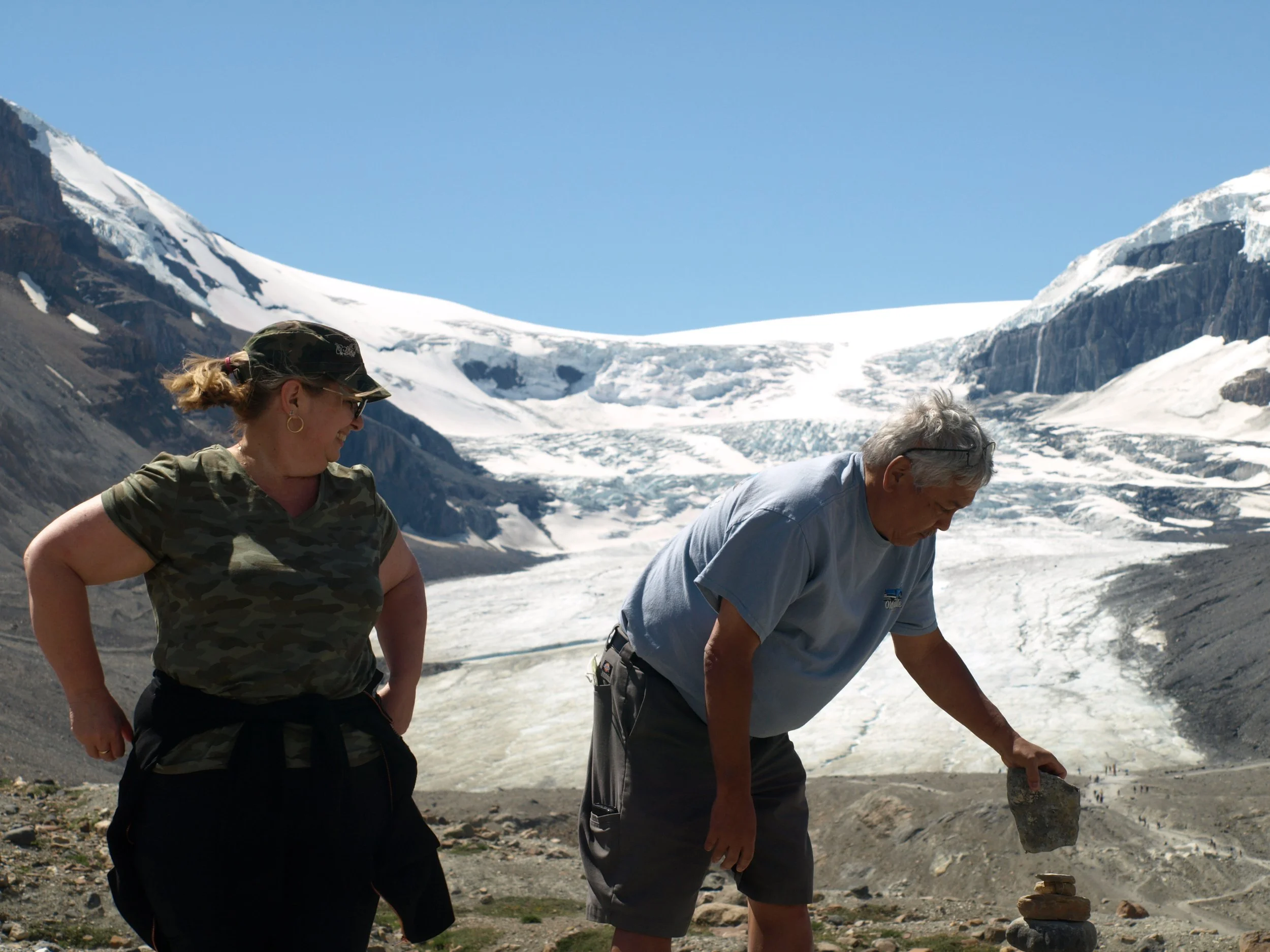 Two people in a mountainous, snowy landscape, one man stacking stones, a woman watching, under a clear blue sky.