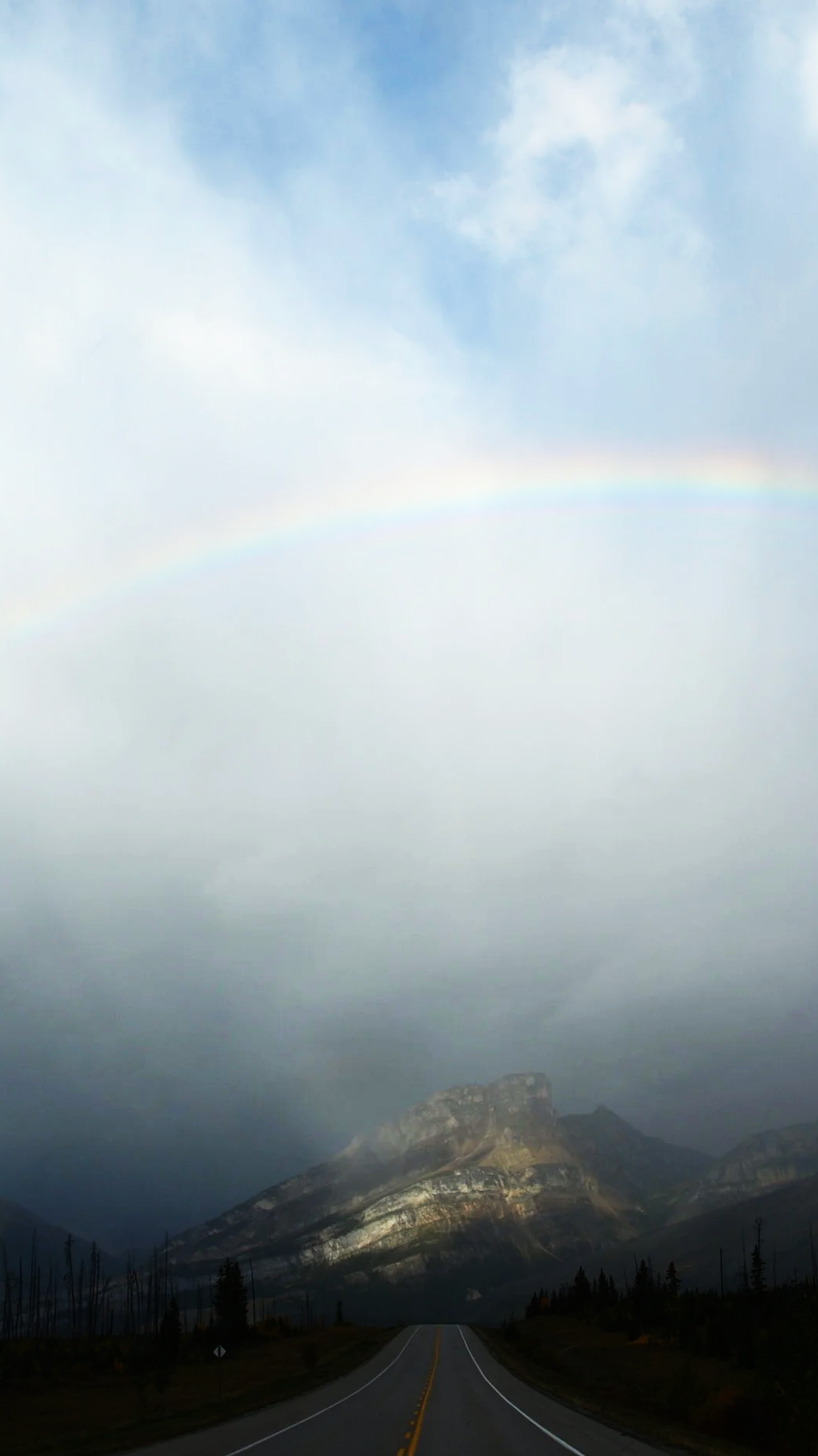A road leading towards a mountain with a rainbow in the sky above, partially covered by clouds.