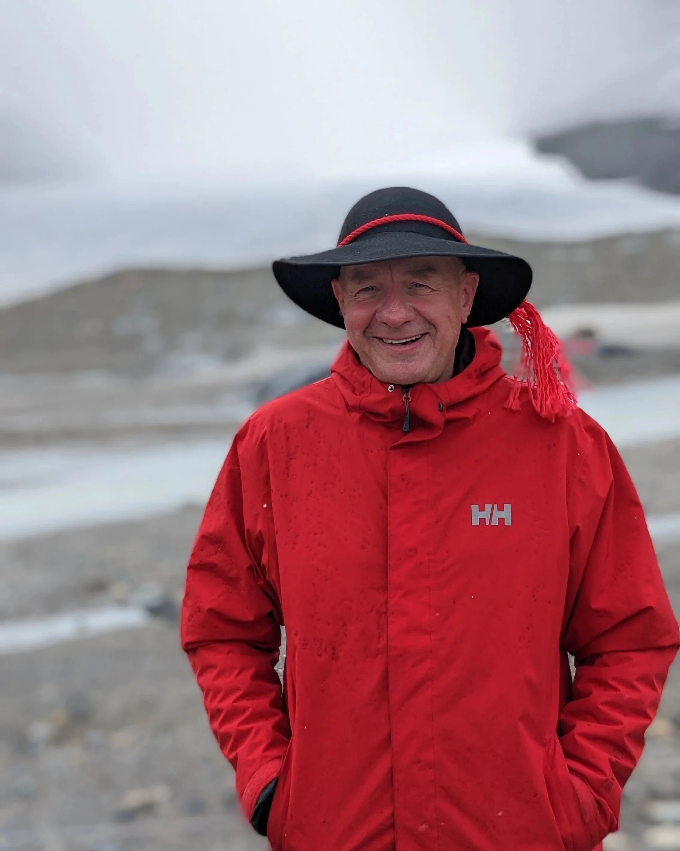 Smiling man in a red jacket and a wide-brimmed black hat outdoors in a cold, mountainous environment.