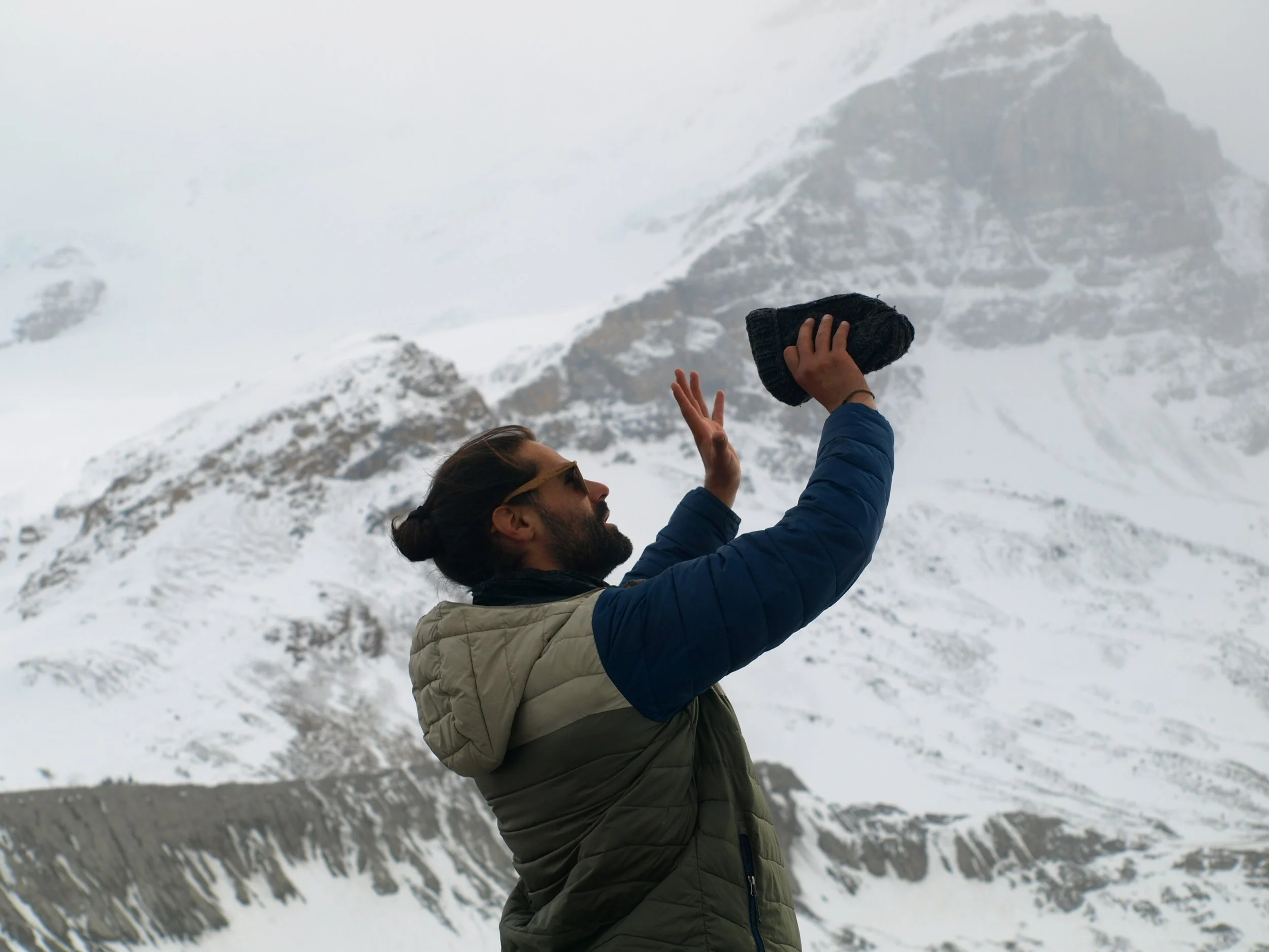 A man with sunglasses, a beard, and a man bun is standing in snowy mountain terrain, holding a black beanie in the air with one hand, possibly taking a selfie or photo.