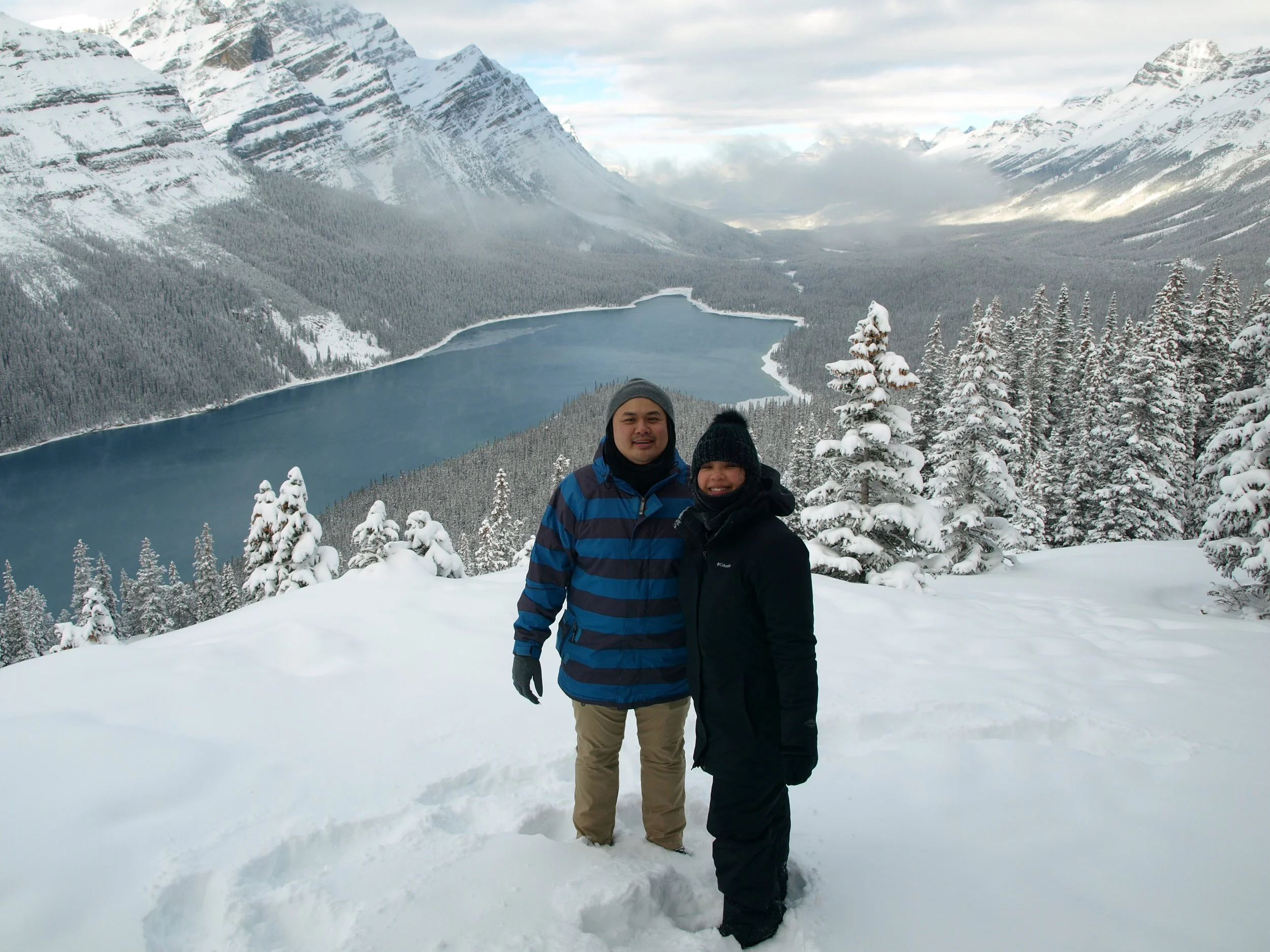 Two people standing in snow-covered landscape with pine trees, a lake, and snow-capped mountains in the background.