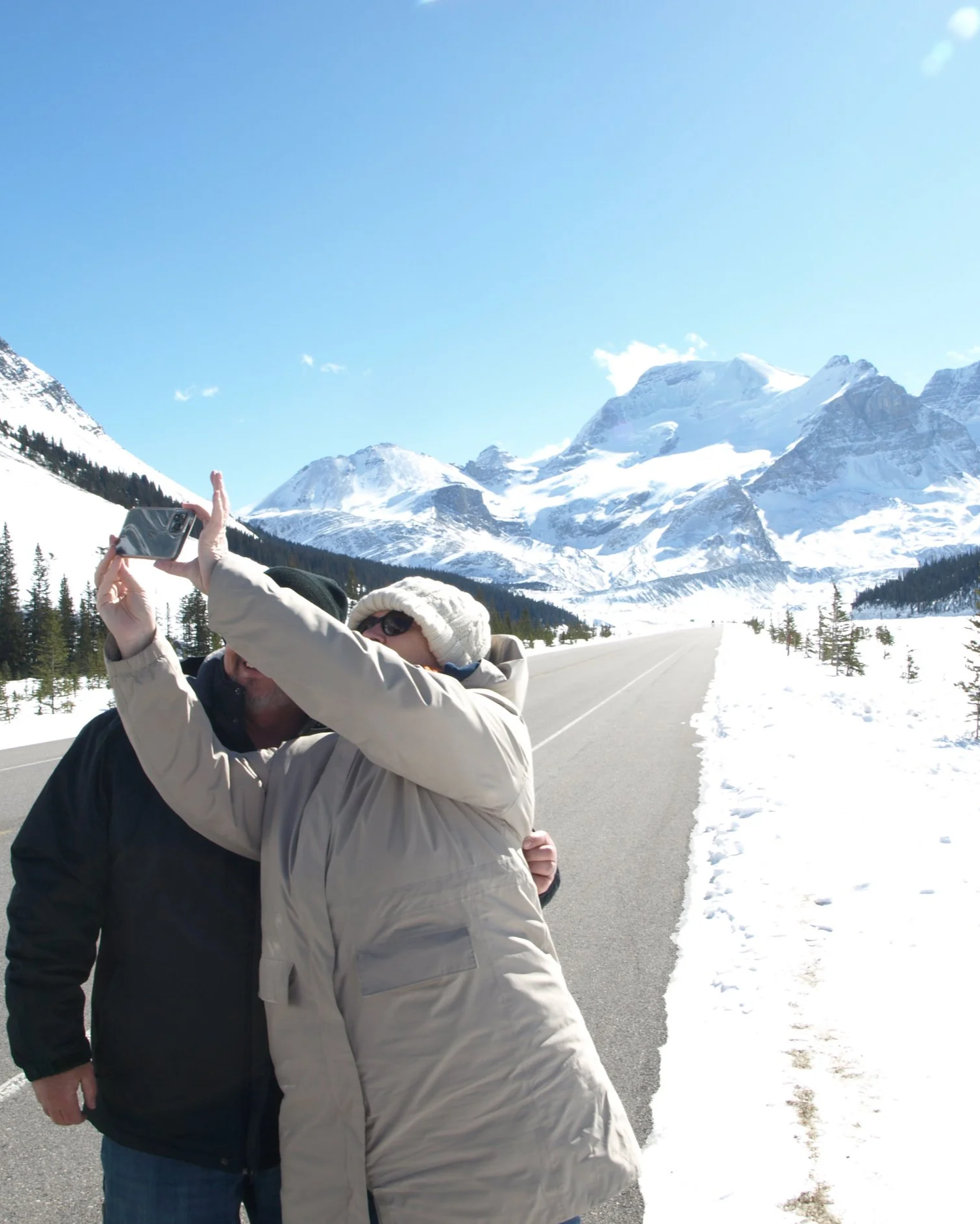 Two people taking a selfie on a snowy mountain road with snow-covered mountains and blue sky in the background.