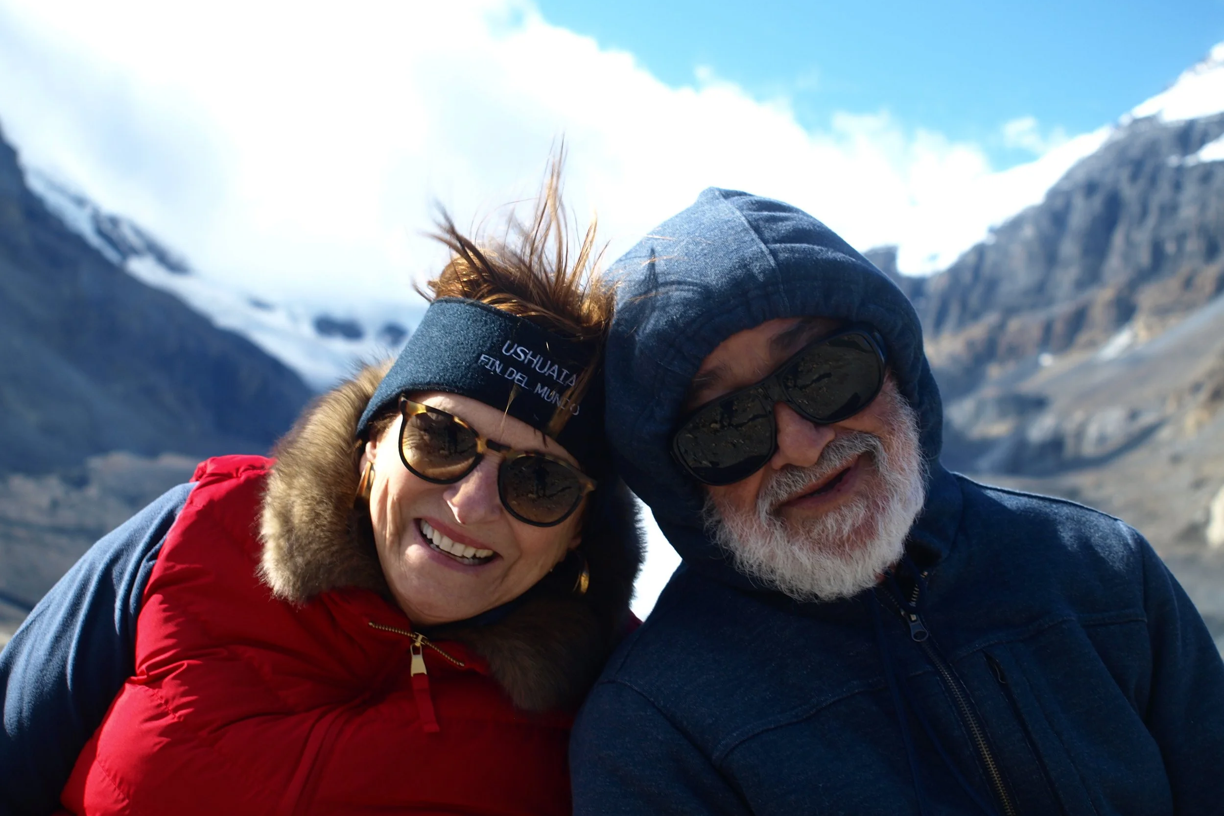 Smiling man and woman in winter clothing and sunglasses outdoors in a mountainous snowy landscape.