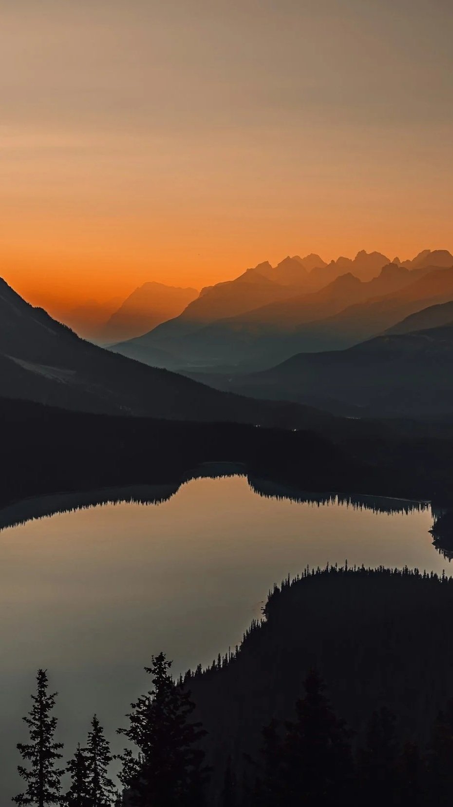 A scenic view of mountains at sunset with a calm lake in the foreground, surrounded by trees.