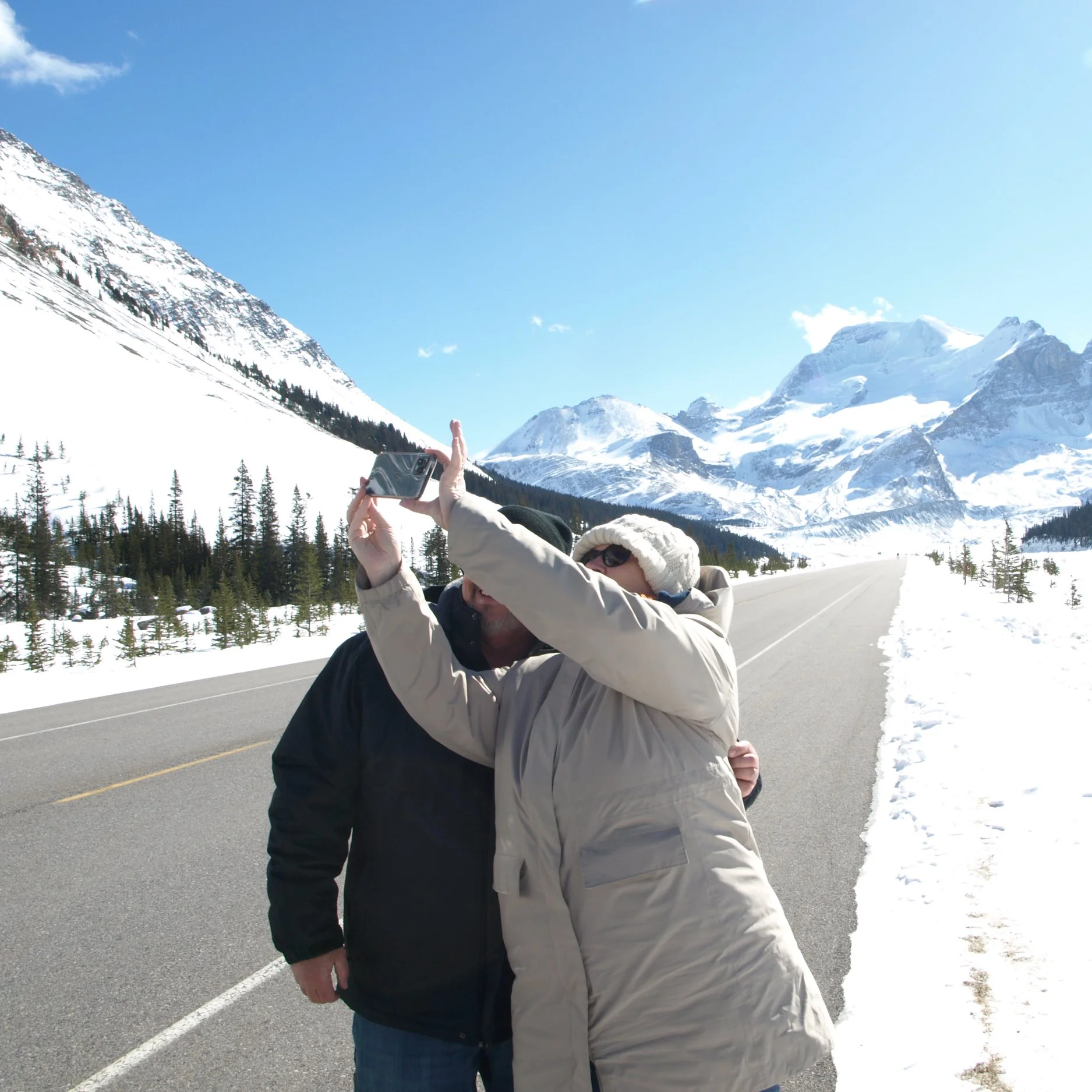 Two people taking a selfie on a snowy mountain road with snow-covered mountains in the background.