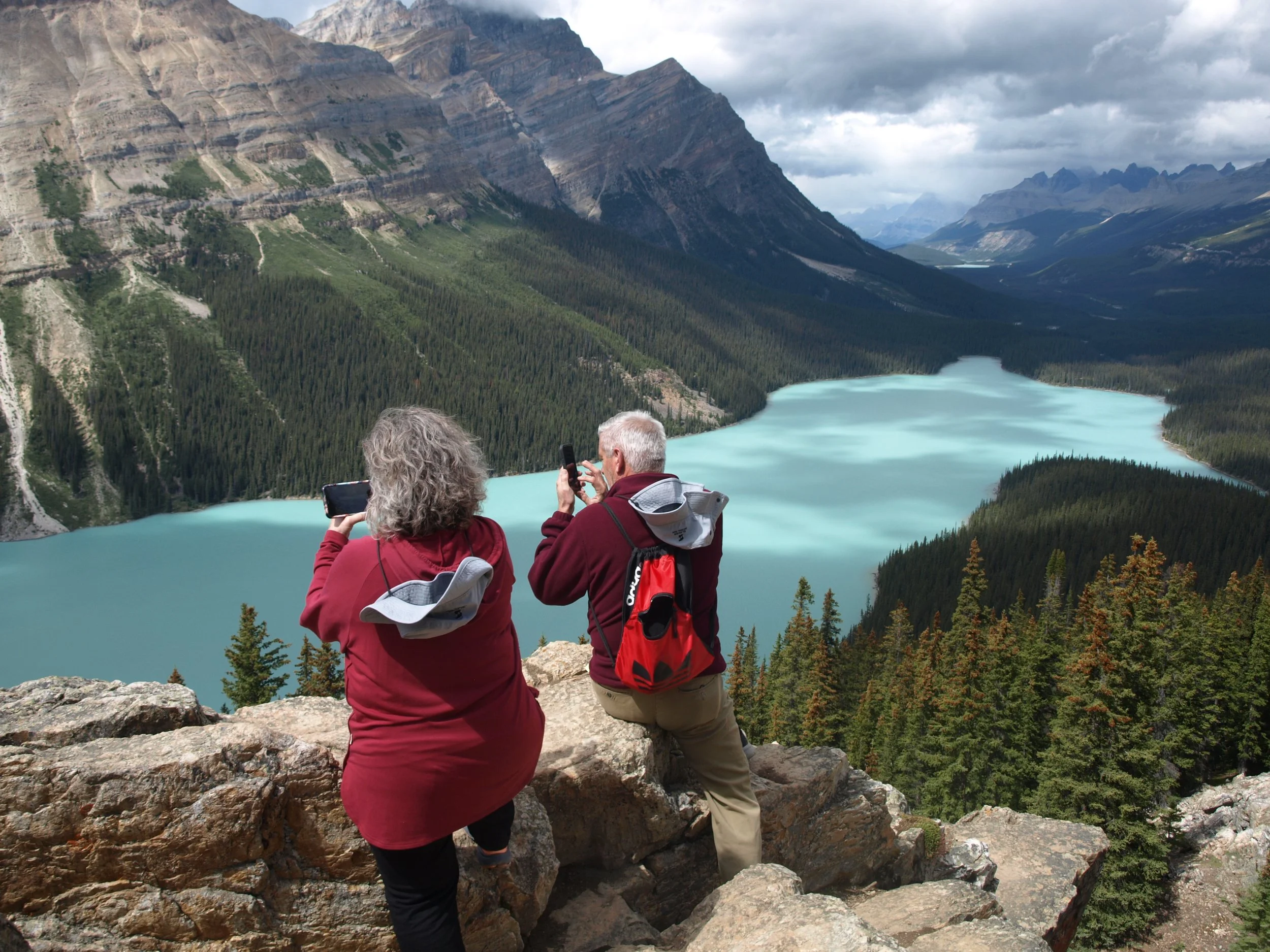 Two tourists, a woman and a man, taking photos of a turquoise lake surrounded by green forest and mountains, from a rocky overlook.