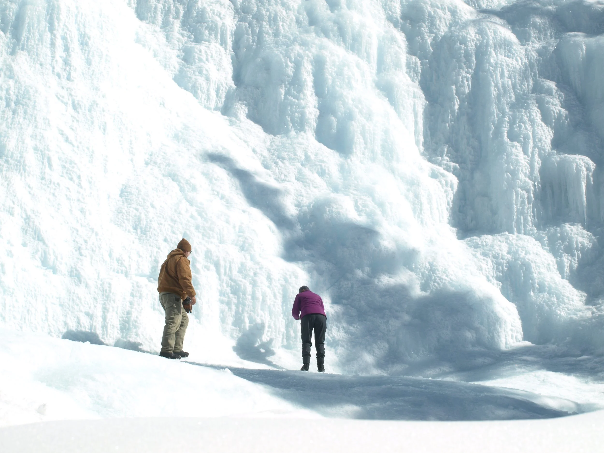 Two people in winter clothing standing on snow, observing a large glacier or ice formation.