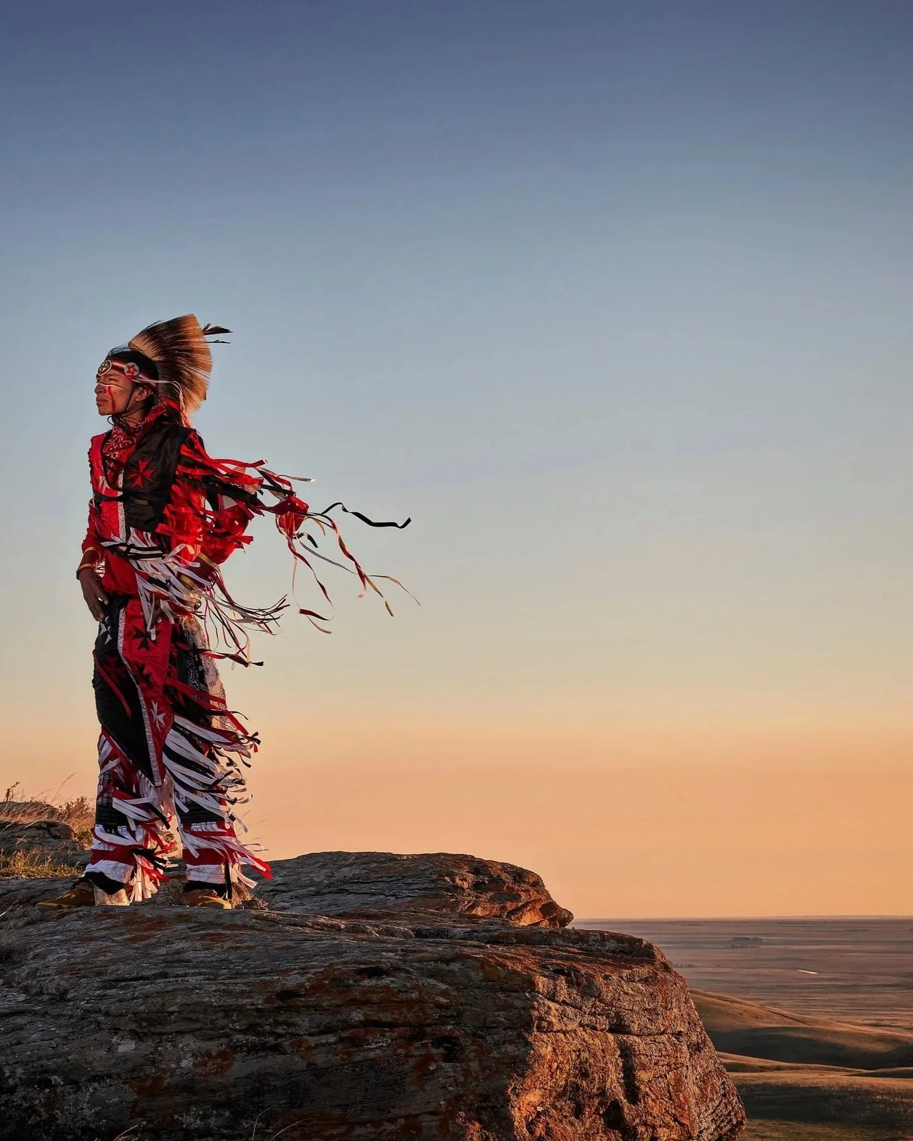 Native American man in traditional colorful regalia, standing on a rock, with a vast landscape and sunset in the background.