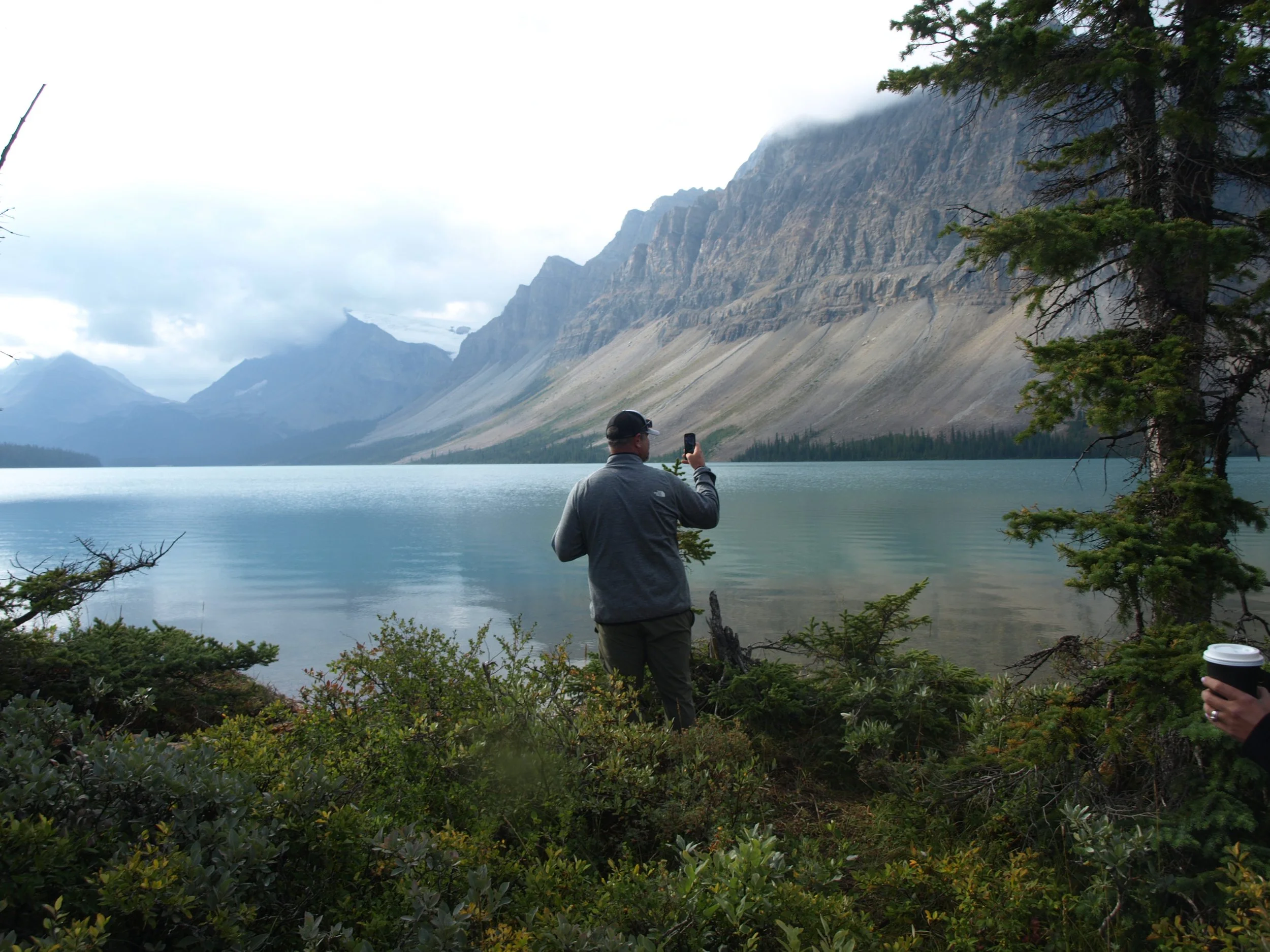 A person taking a photo of a mountain lake with a smartphone, standing among green bushes and trees in a mountainous landscape with cloudy skies.