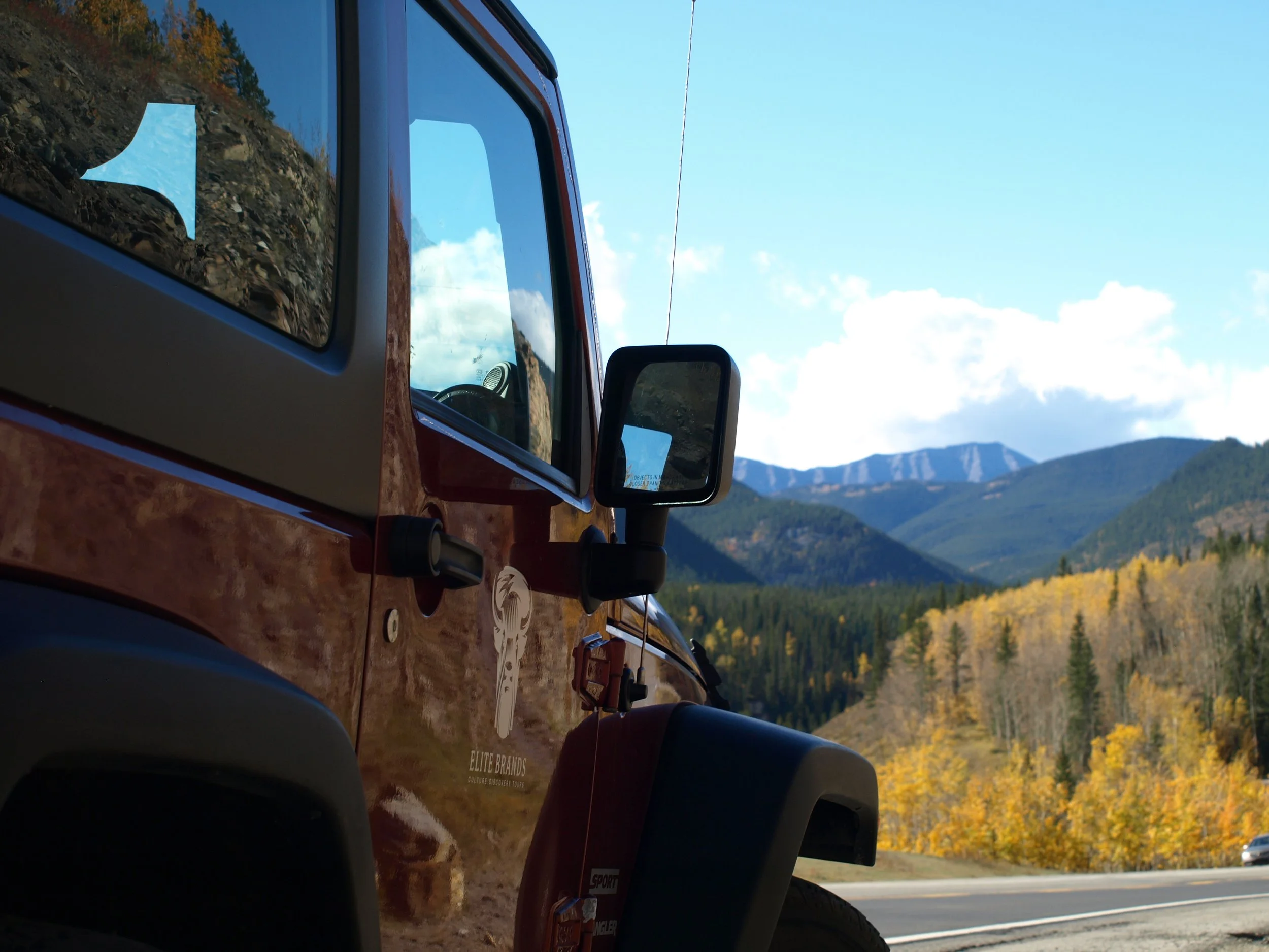 Close-up of a red Jeep with a sticker on the door, parked by a mountain road with a scenic view of mountains and trees in fall colors.