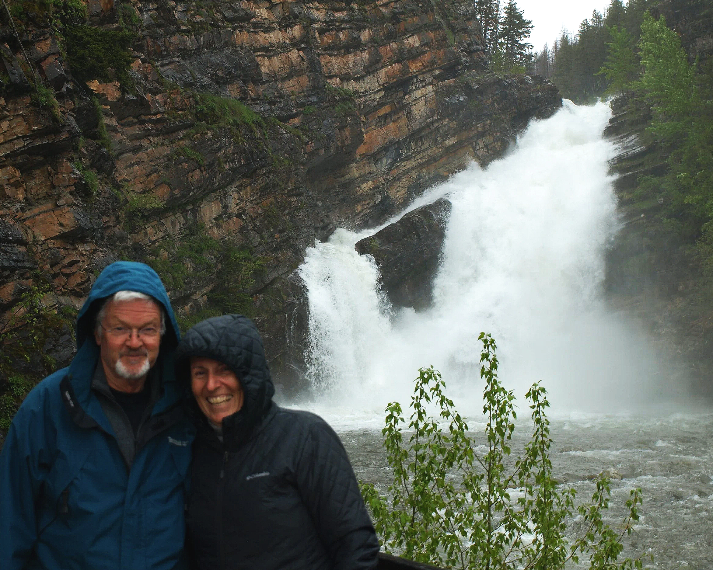 Two people in rain jackets standing in front of a waterfall in a rocky, forested area.