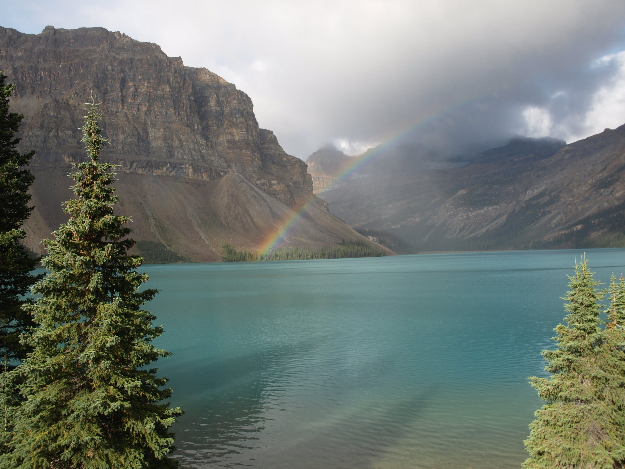Scenic view of a turquoise lake with evergreen trees in the foreground, mountains in the background, and a rainbow arching over the mountains on a cloudy day.
