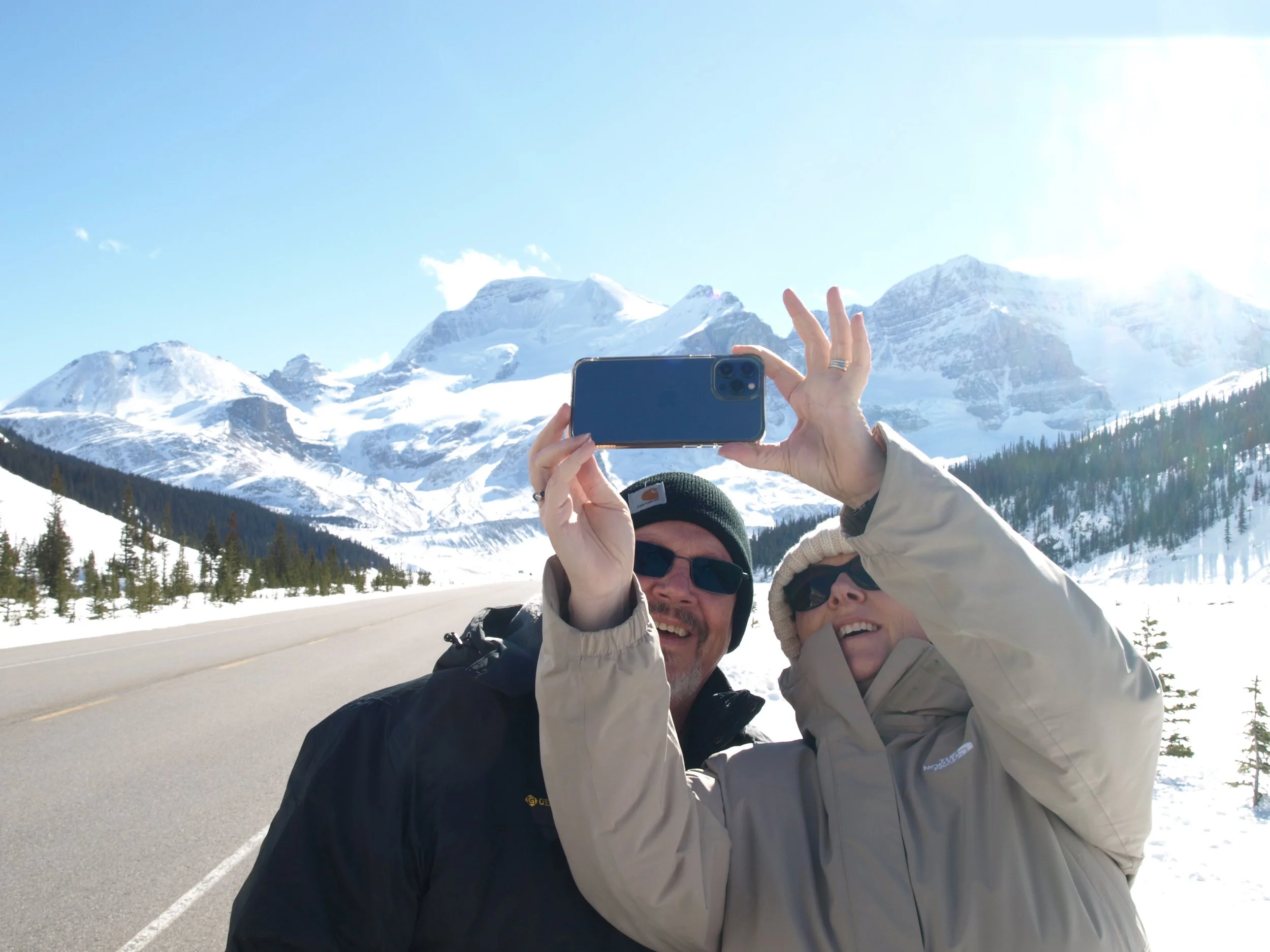 Two people taking a selfie in front of snow-covered mountains and trees.