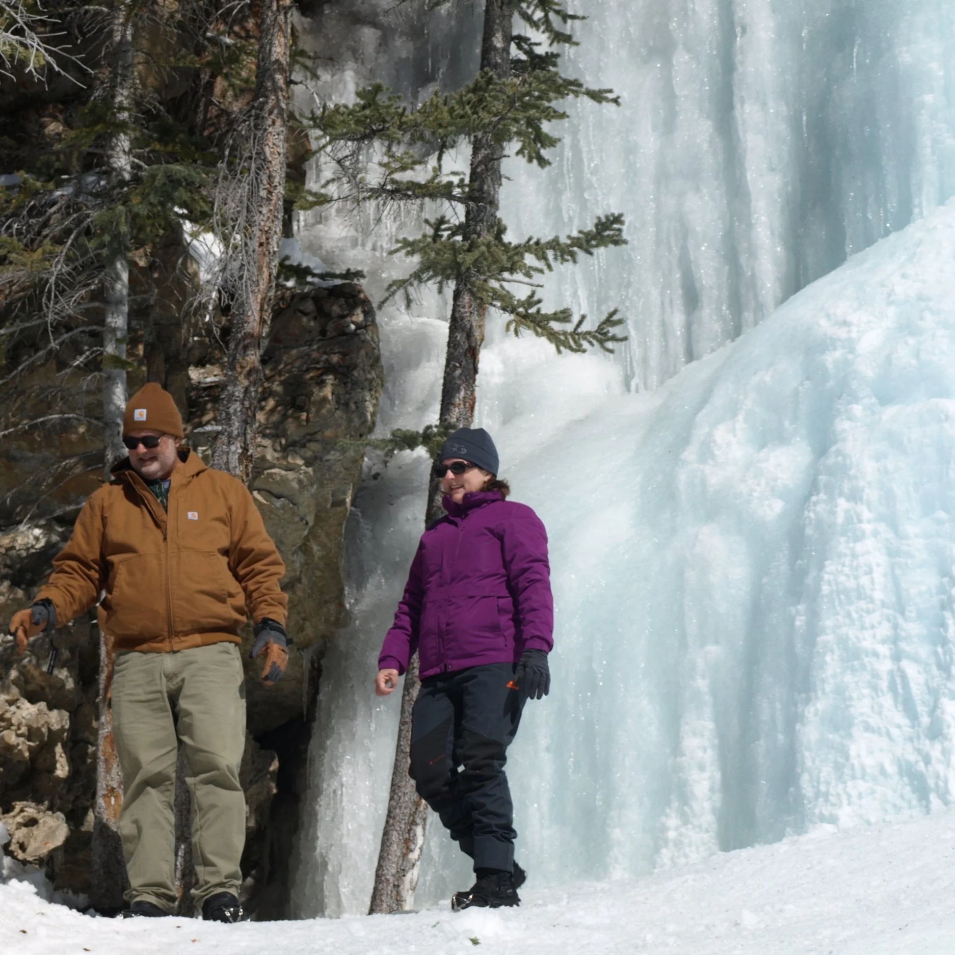 Two people in winter clothing standing near a frozen waterfall and snow-covered terrain, with evergreen trees and rock formations in the background.