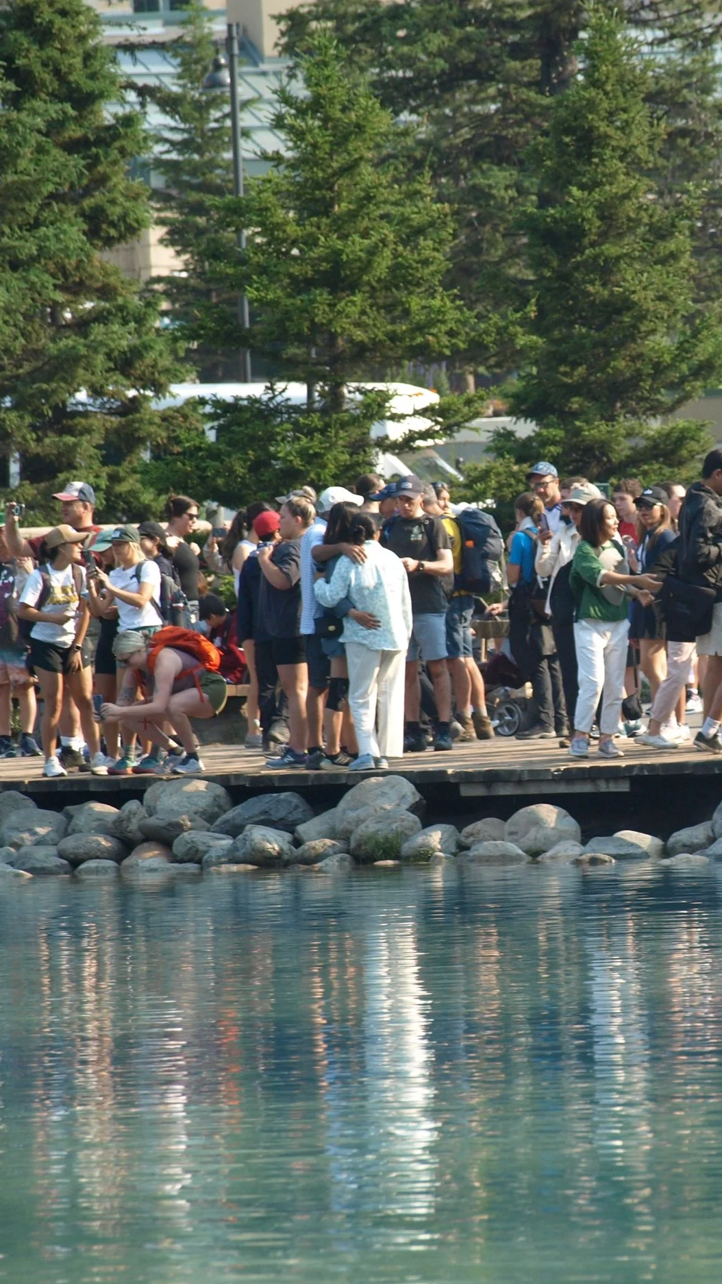 A large crowd of people gathering on a wooden dock by a body of water with trees and buildings in the background.