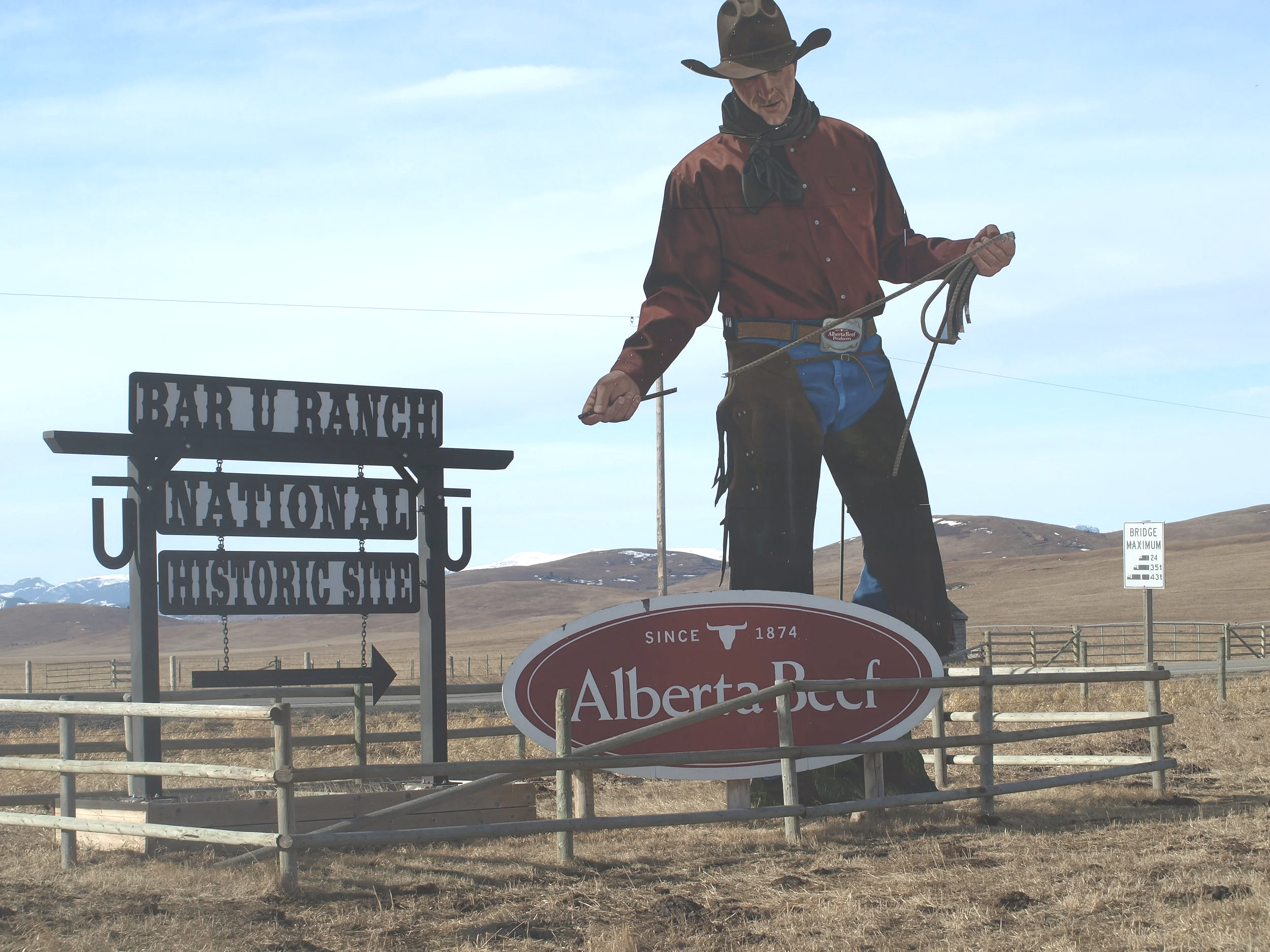 A cowboy statue holding a rope in front of Alberta Beef and Bar U Ranch National Historic Site signs in a rural landscape.