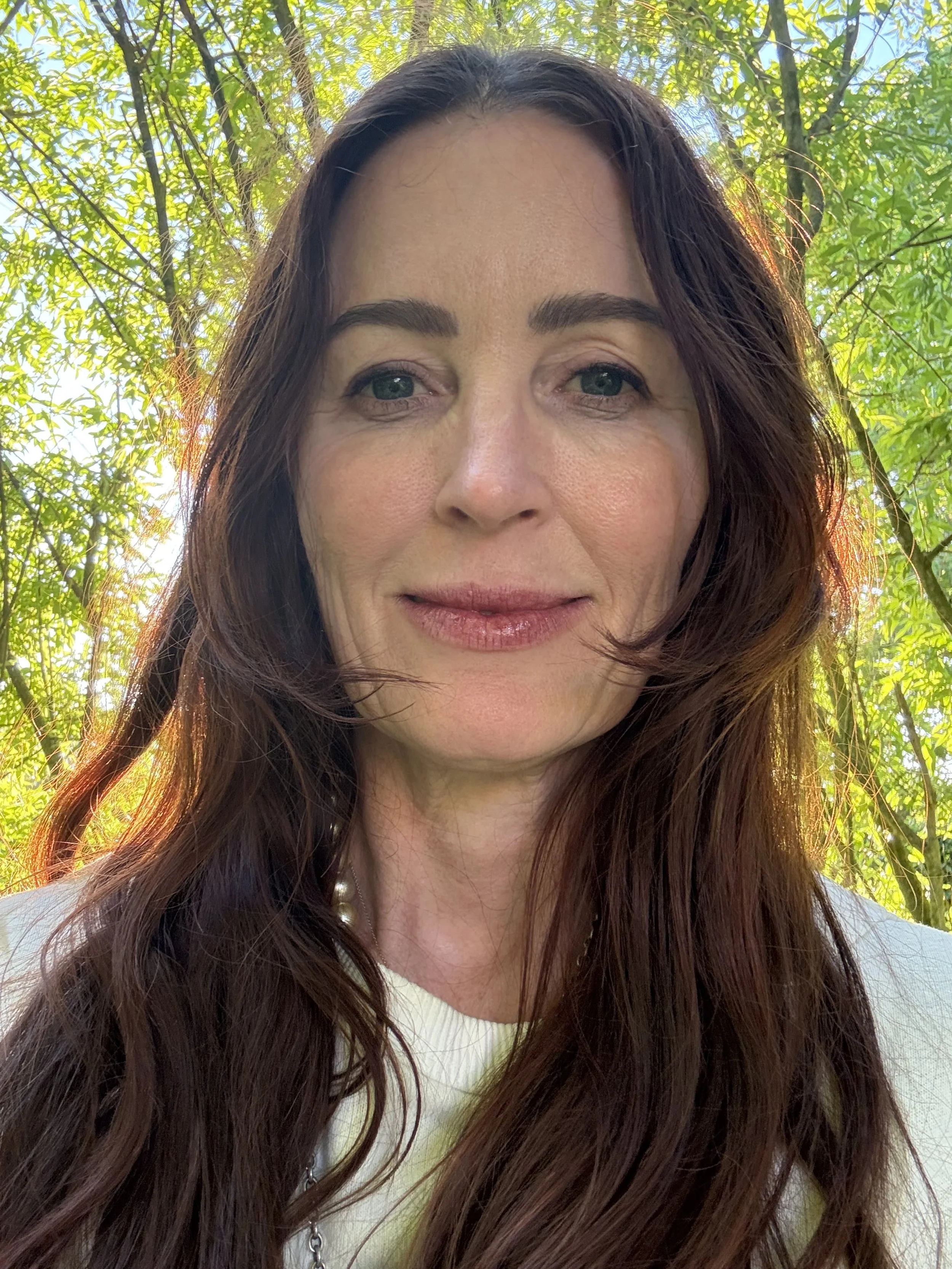 A woman with long brown hair is outdoors in a green leafy area, smiling softly at the camera.