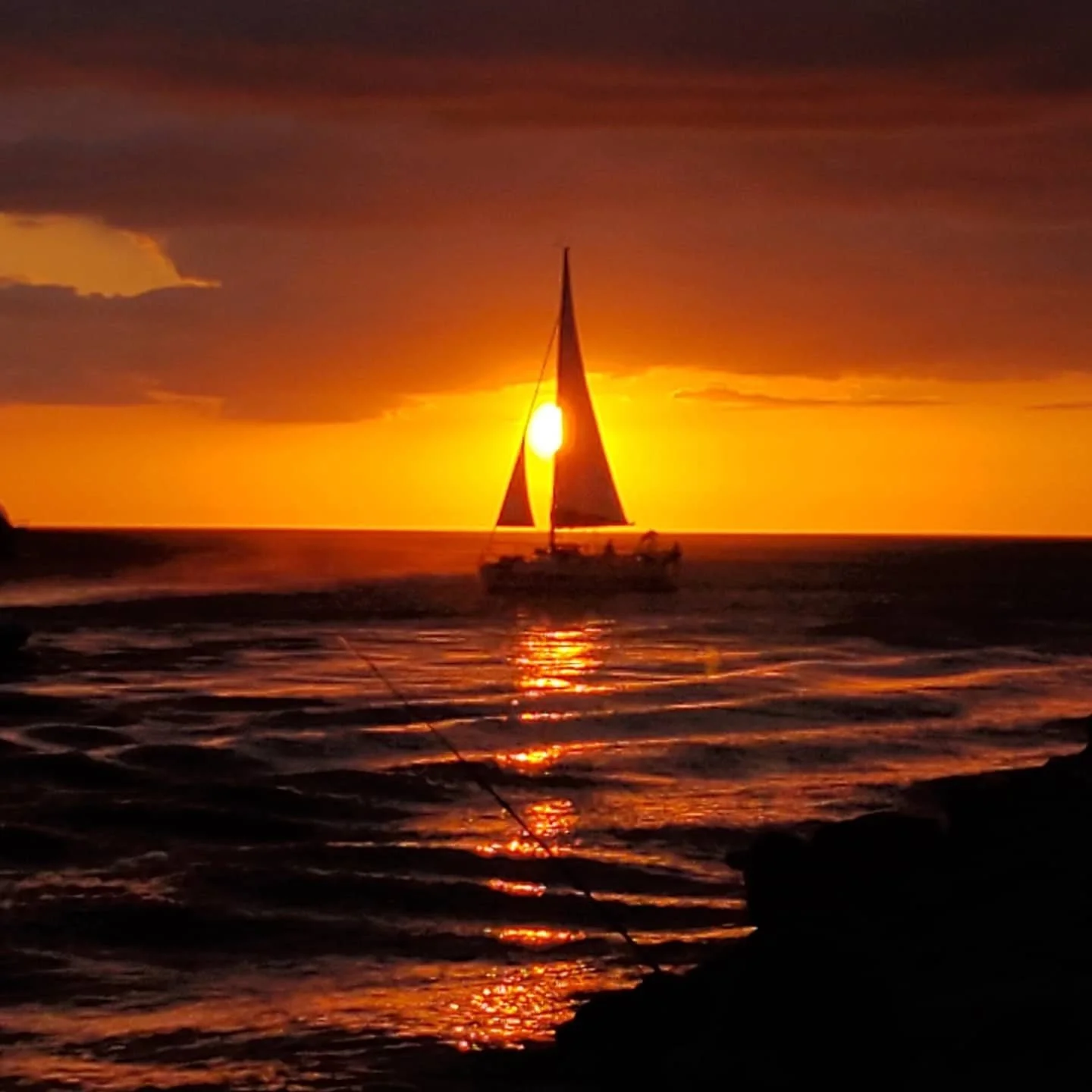 A sailboat on the ocean during sunset with the sun aligned behind the sail, casting a golden reflection on the water.