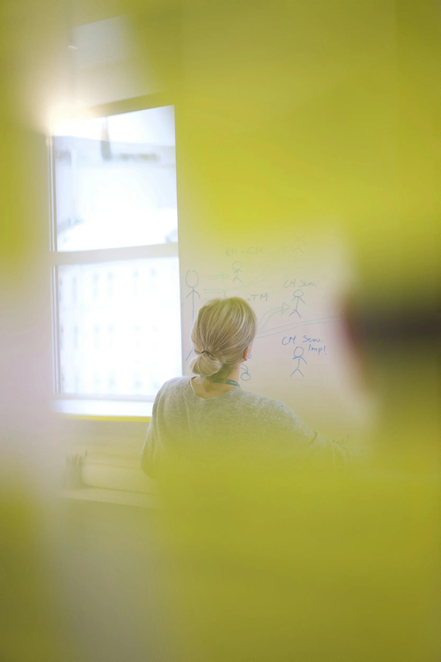 A person with blonde hair tied in a low bun, seen from behind, leading a workshop in a room with a large window. The person is standing near a whiteboard with sketches and notes, with a cityscape visible through the window.